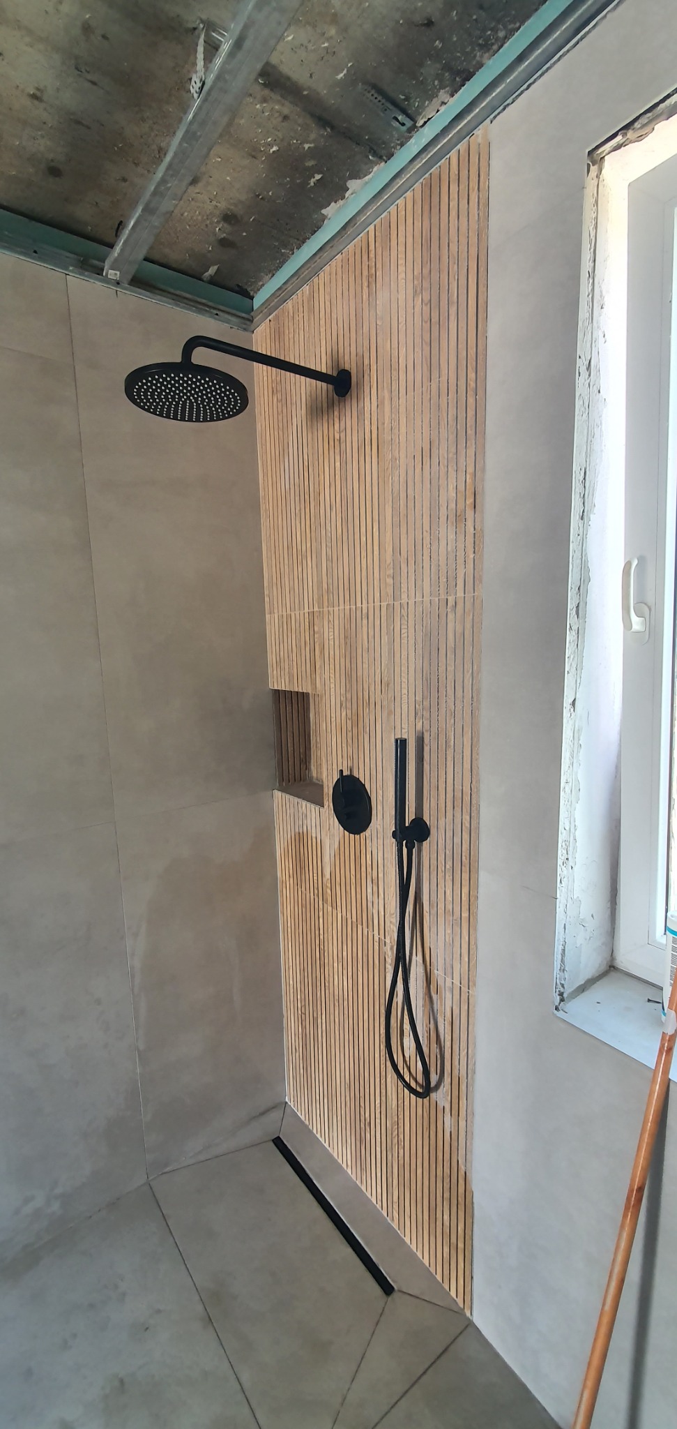 Nowoczesny prysznic z deszczownicą w kolorze czarnym, drewnianą ścianką z wnęką i odpływem liniowym. Widoczne niedokończone wykończenie sufitu i okna.