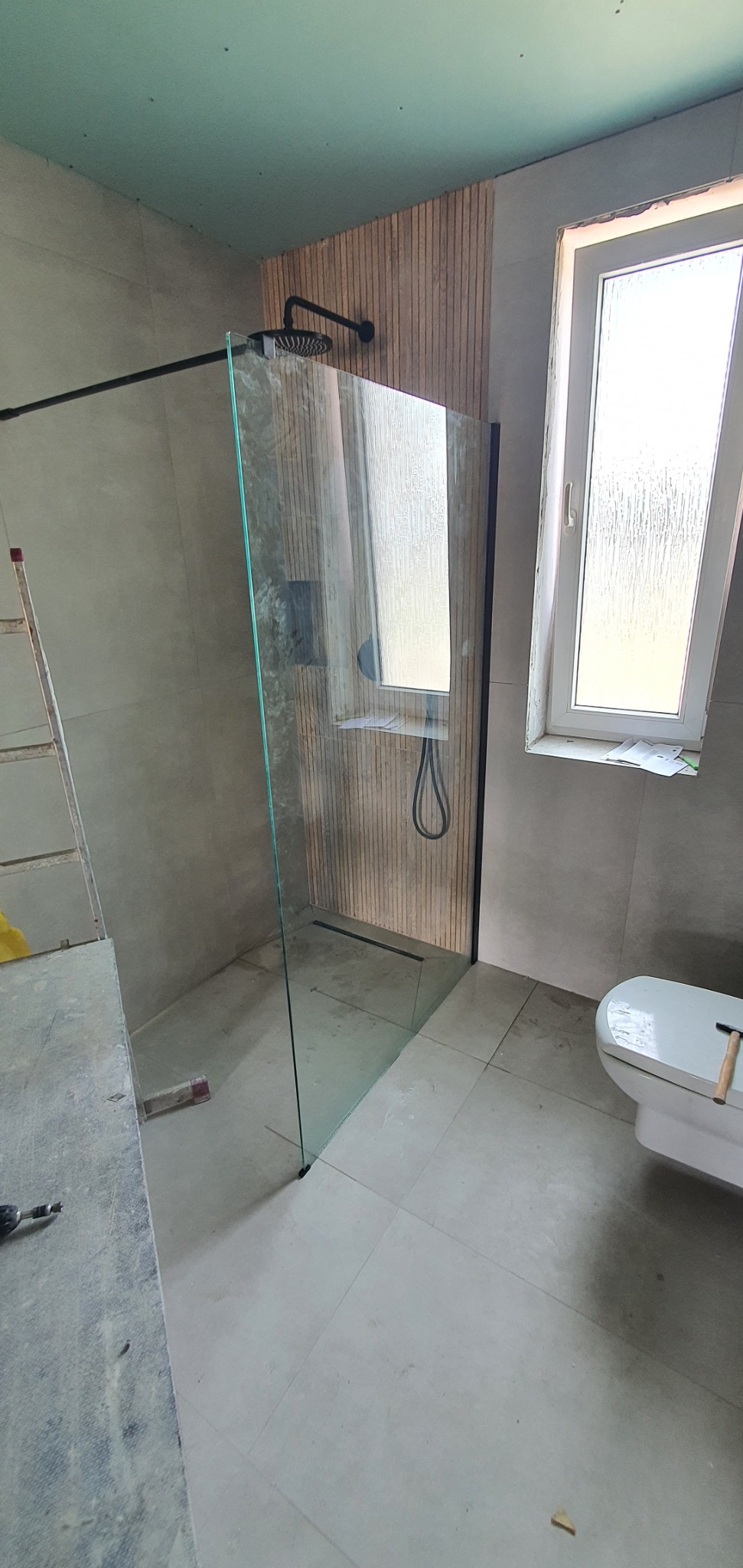 Nowoczesna kabina prysznicowa typu walk-in z czarnymi detalami, zamontowana w łazience wykończonej płytkami w odcieniach szarości i drewnianymi panelami. Widoczny fragment toalety.