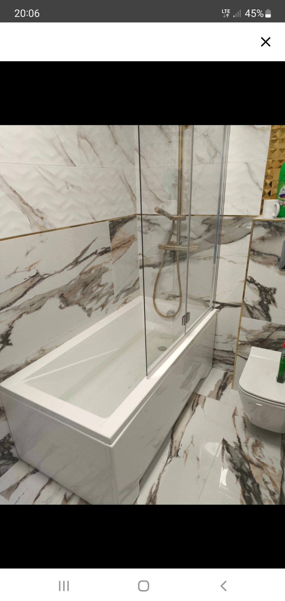 Biała wanna z parawanem nawannowym i złotą armaturą na tle marmurowych płytek w łazience. Widoczny fragment toalety i środki czystości.