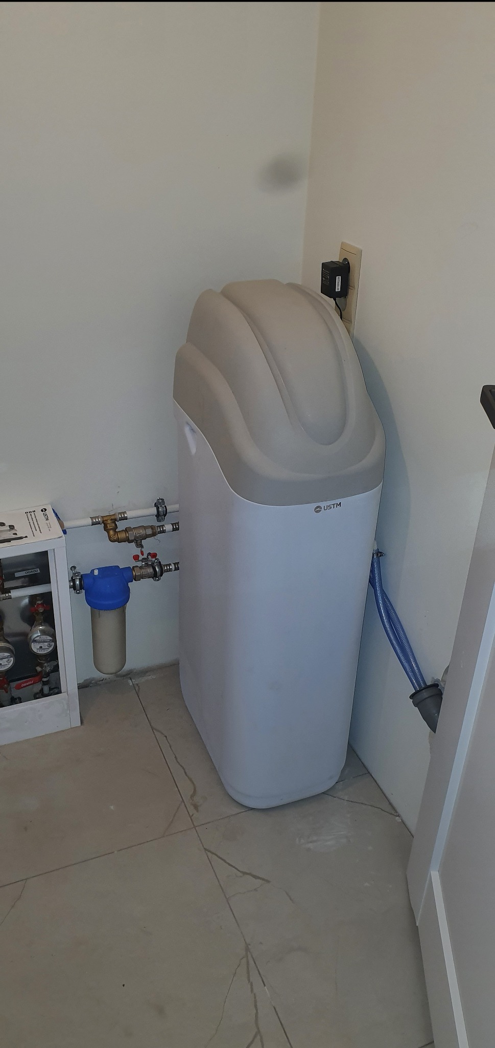 Biały zmiękczacz wody USTM, z niebieskim wężem odpływowym, podłączony do instalacji wodnej. Widoczny filtr narurowy i rozdzielacz. Białe ściany i płytki podłogowe.