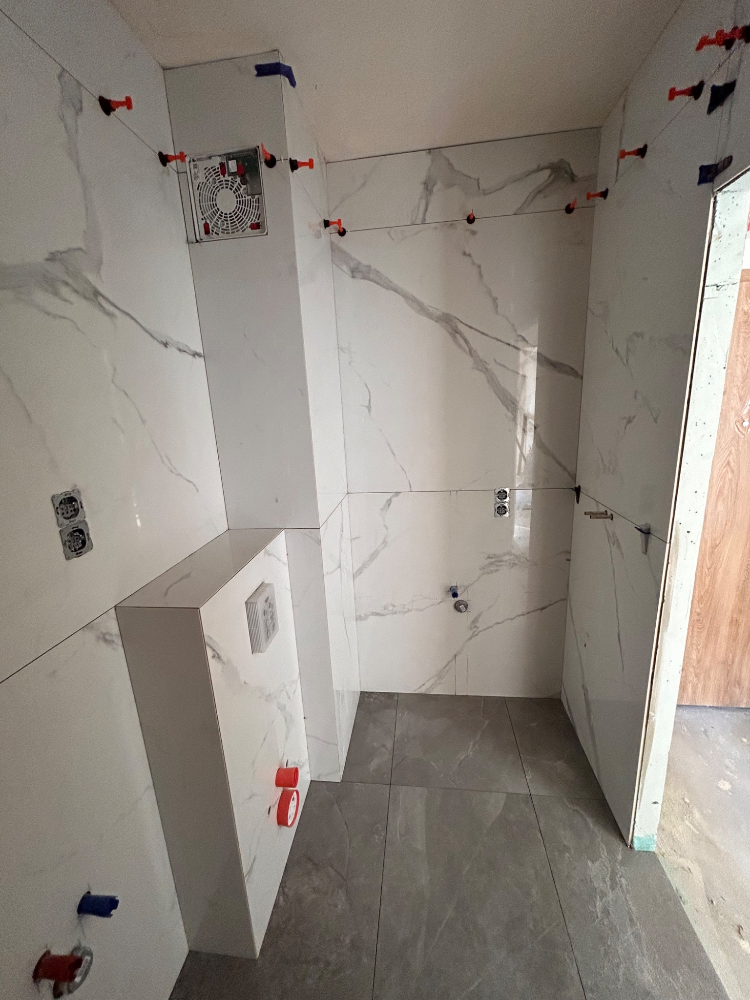 Łazienka w trakcie układania płytek imitujących marmur, widoczne systemy poziomowania płytek, zamontowany stelaż podtynkowy WC, szare płytki podłogowe.