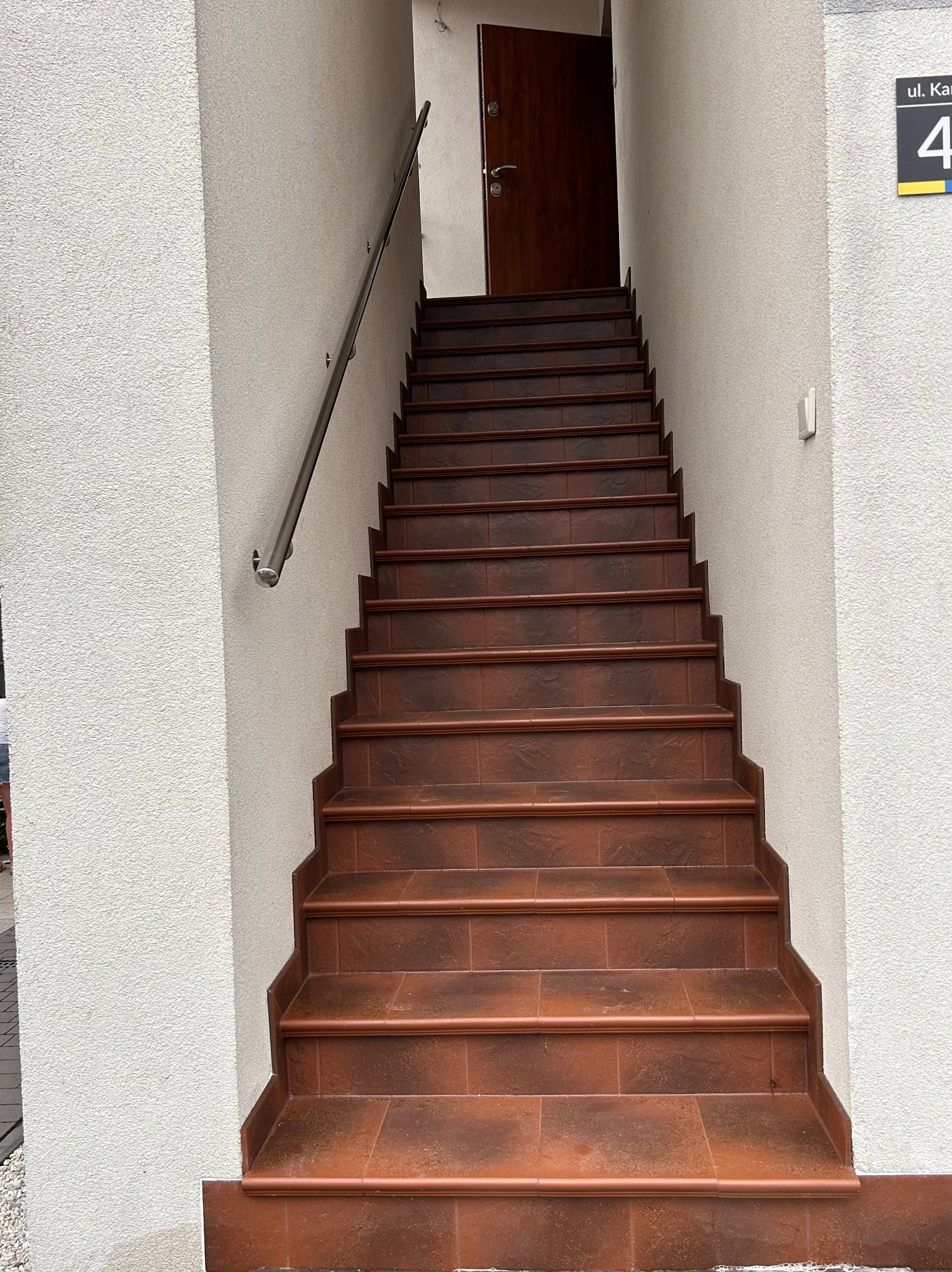 Zewnętrzne schody wejściowe wyłożone brązową terakotą, z metalową poręczą po lewej stronie i numerem domu '4' po prawej.