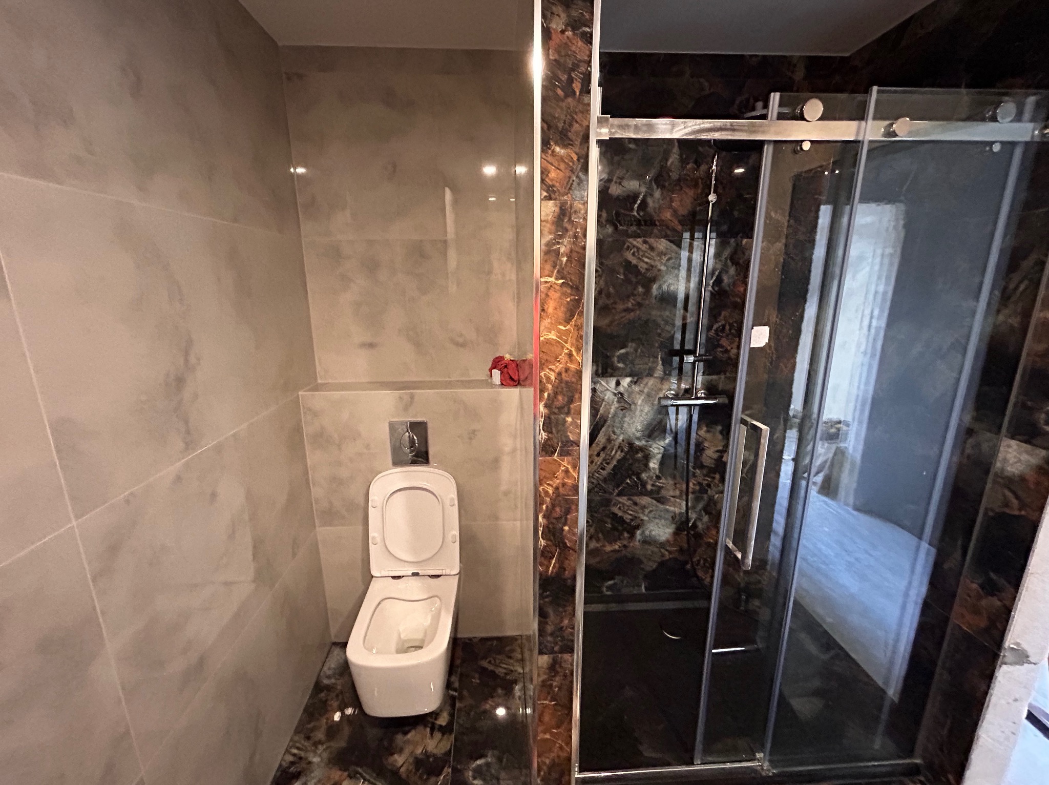 Wykończona łazienka z toaletą z otwartą klapą i kabiną prysznicową z czarnymi marmurkowymi płytkami, jasne płytki na ścianach, widoczny fragment czerwonego ręcznika.