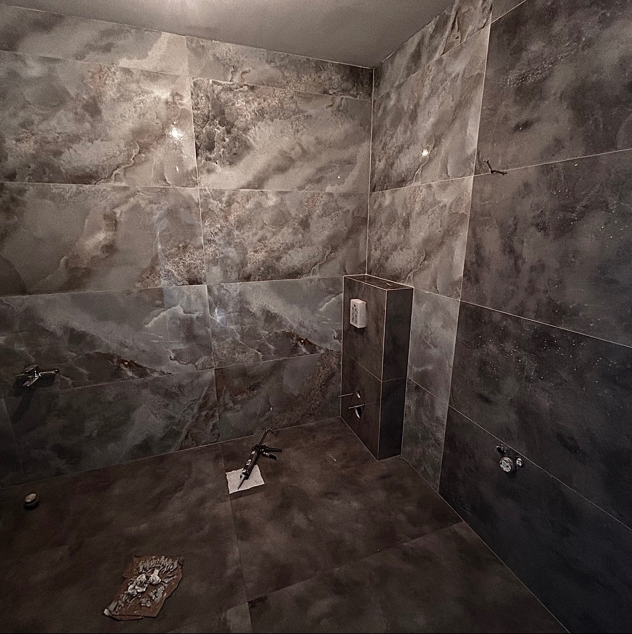 Łazienka w trakcie wykańczania, ściany i podłoga wyłożone płytkami imitującymi marmur w odcieniach szarości, widoczna puszka podtynkowa, narzędzia glazurnicze i fragment uszkodzonej płytki...