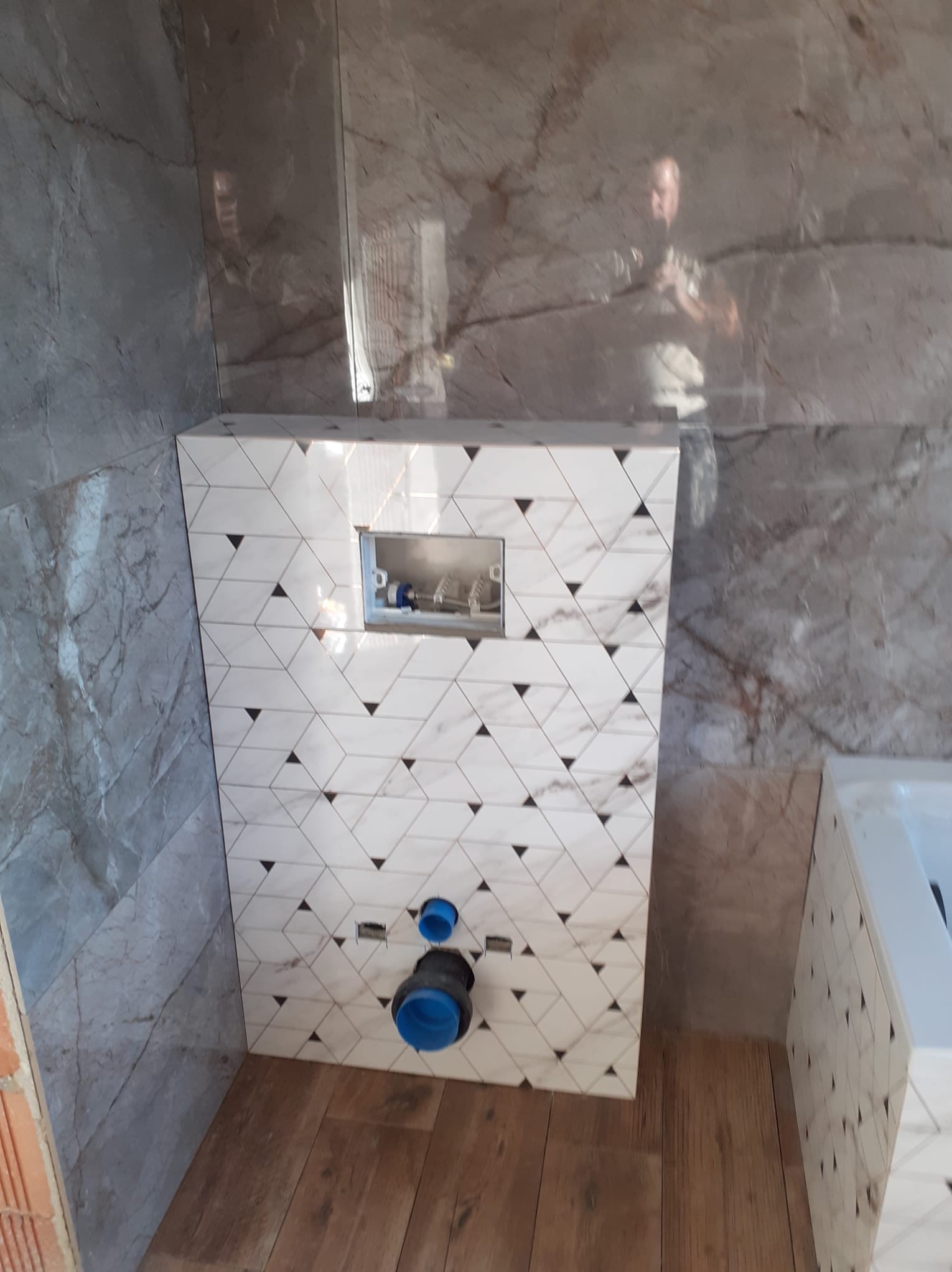 Ścianka zabudowy stelaża WC wykończona biało-czarną mozaiką trójkątną, na ścianach i podłodze płytki imitujące marmur, widoczne niebieskie elementy instalacji sanitarnej.