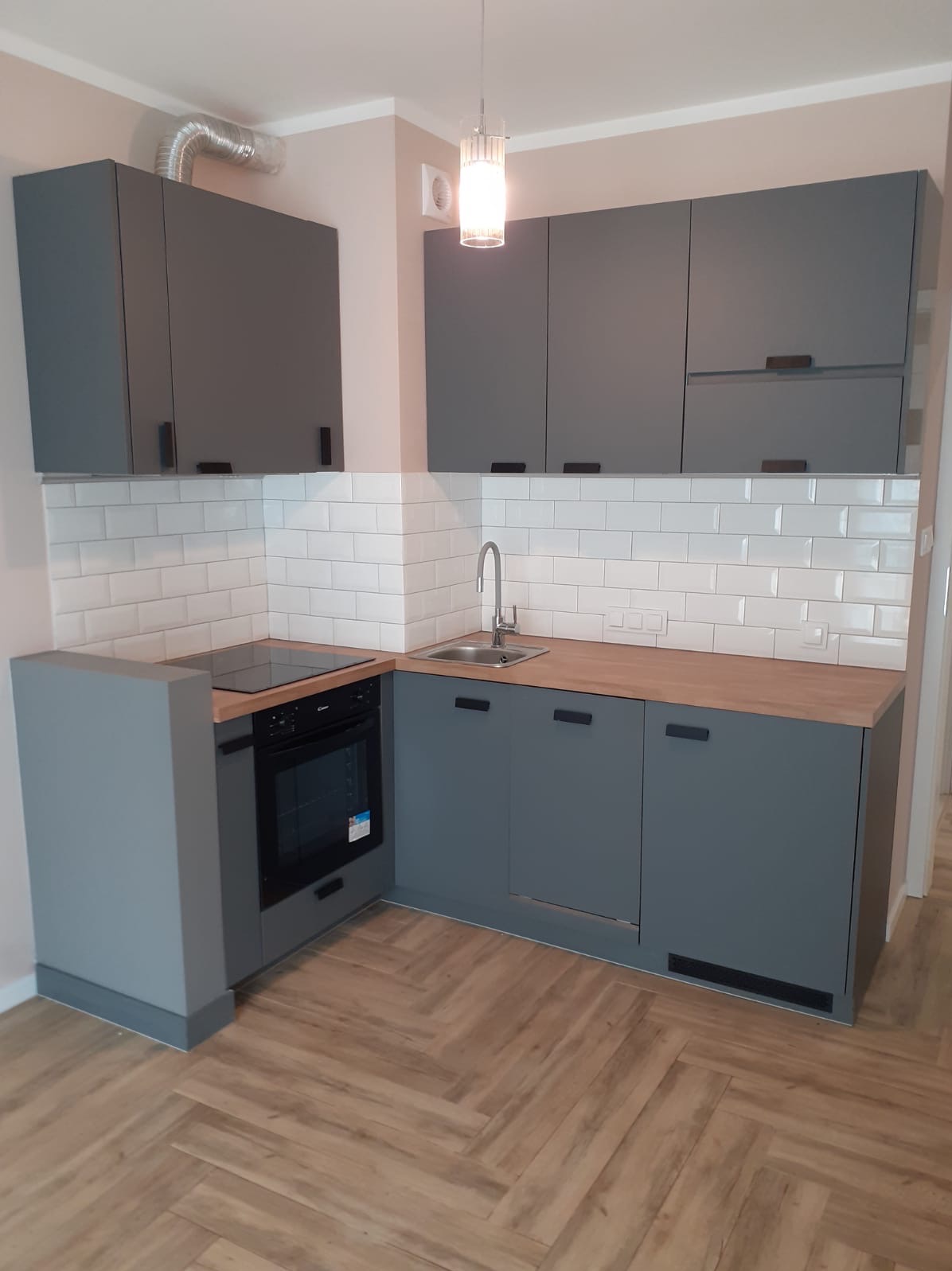 Szara zabudowa kuchenna z drewnianym blatem i białymi płytkami ułożonymi w cegiełkę, widok na fragment kuchni z parkietem w jodełkę.