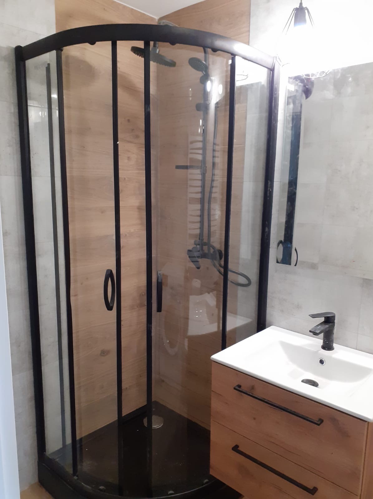 Nowoczesna kabina prysznicowa z czarną ramą, drewnopodobne płytki na ścianach wewnątrz kabiny, umywalka z szafką z drewnianym frontem, czarna bateria.