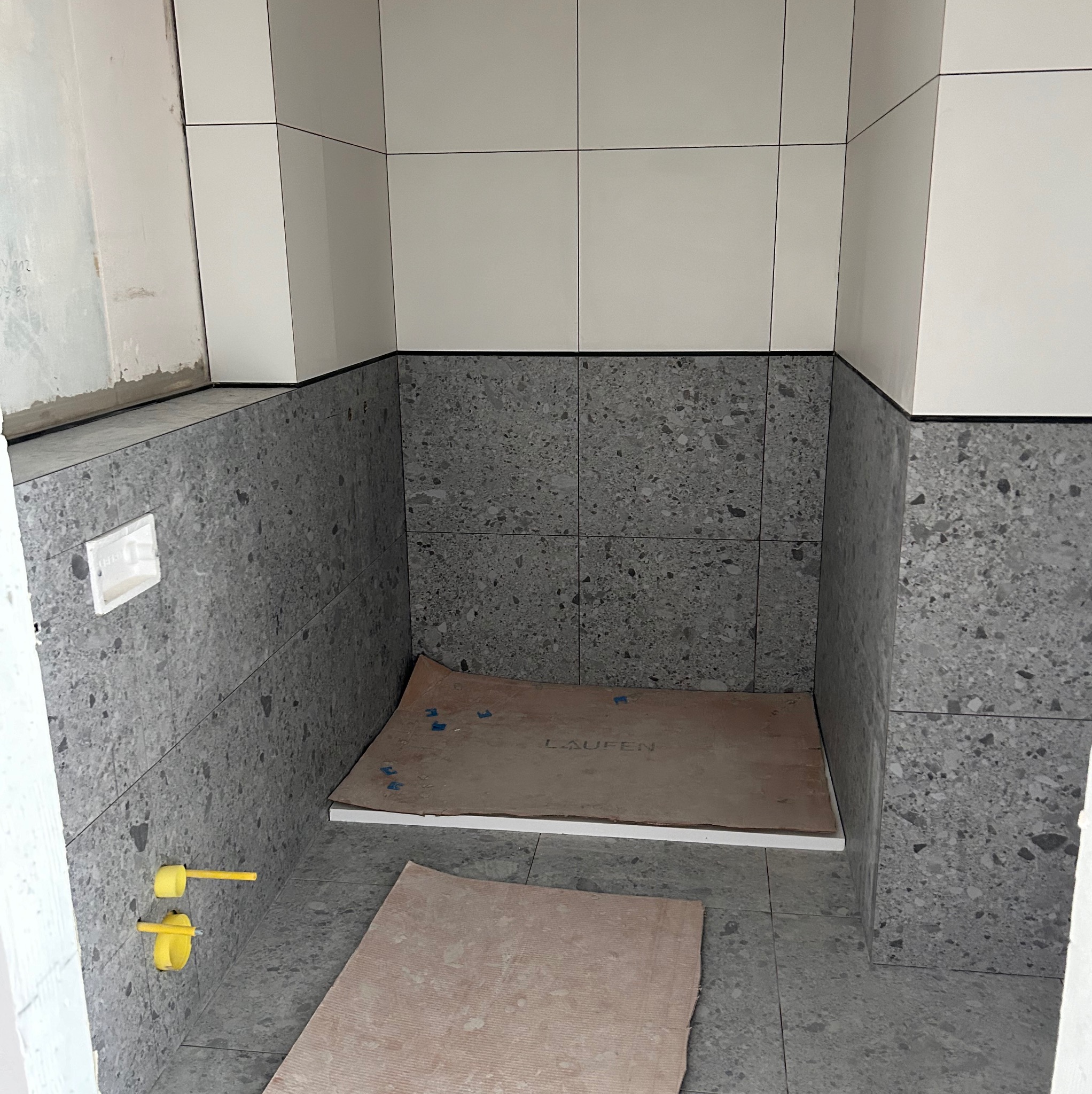 Wnęka prysznicowa w trakcie wykańczania, z częściowo położonymi białymi i szarymi płytkami, widoczne żółte przyłącza wodne i zabezpieczone podłoże pod brodzik z napisem LAUFEN.
