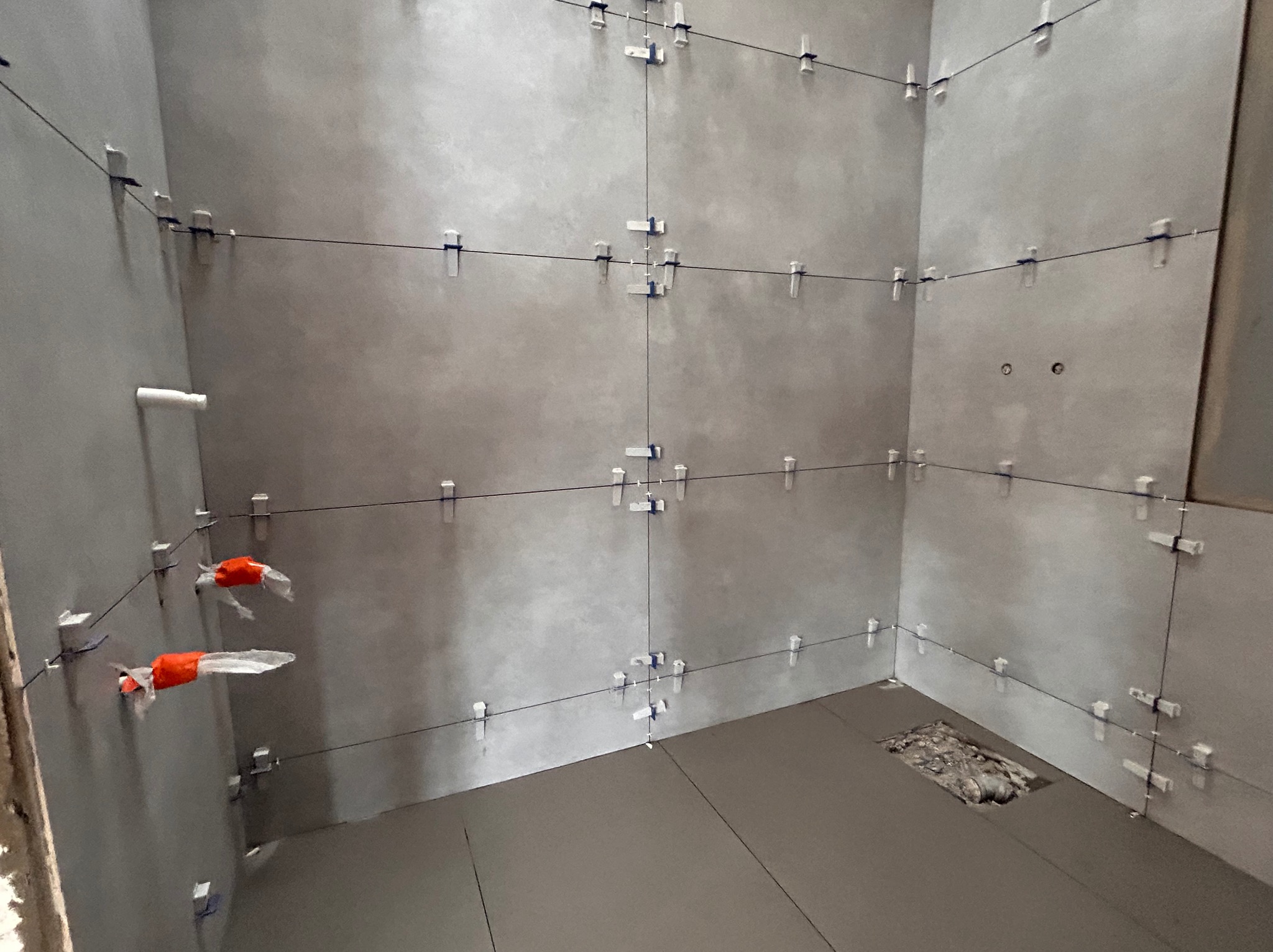 Szare płytki wielkoformatowe na ścianach łazienki w trakcie montażu, z użyciem systemu poziomowania z klipsami i sznurkami, widoczny otwór w podłodze.