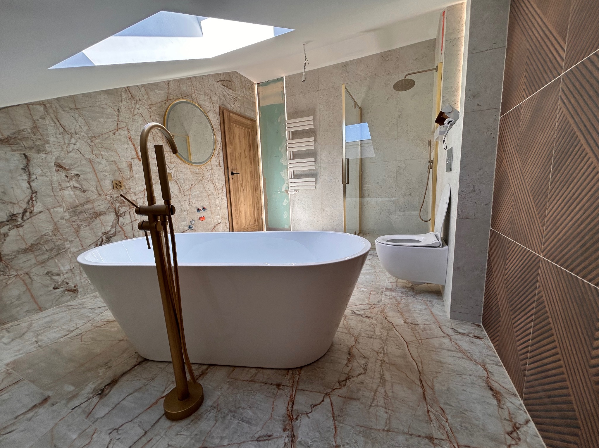 Elegancka łazienka z wanną wolnostojącą na marmurowej podłodze, kabiną prysznicową ze złotymi detalami i oknem w suficie.