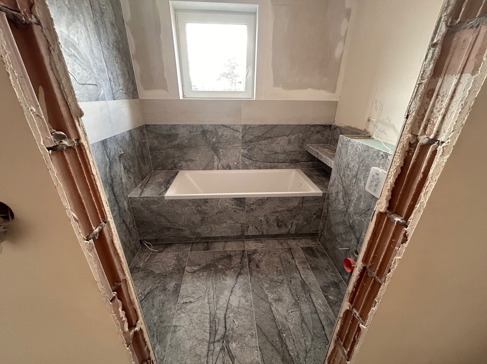 Biała wanna w łazience z szarymi płytkami marmurkowymi, widok przez surowy otwór drzwiowy. Okno w tle. Częściowo wykończone ściany.