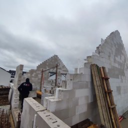 Volodymyr Hapun - Wznoszenie ścian domu z bloczków betonu komórkowego, widoczny pracownik w czarnej bluzie z napisem 'XXIV', szalunki i zbrojenia w tle, pochmurne niebo.