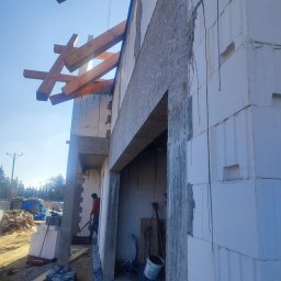 Volodymyr Hapun - Wznoszenie ścian domu z bloczków betonu komórkowego, widoczne drewniane elementy konstrukcyjne dachu i pracownik w czerwonej kurtce.