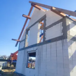 Wznoszenie ścian domu z białych bloczków, widoczne drewniane elementy konstrukcji dachu oraz dźwig w tle.