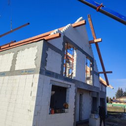 Volodymyr Hapun - Wznoszenie dwukondygnacyjnego domu z białych bloczków, widoczny dźwig podający drewniane elementy konstrukcji dachu, na pierwszym planie osoba przeglądająca dokumenty.