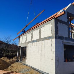 Volodymyr Hapun - Wznoszenie ścian domu z betonu komórkowego; dźwig podnosi drewniany element konstrukcji dachu, widoczna izolacja przeciwwilgociowa fundamentów.