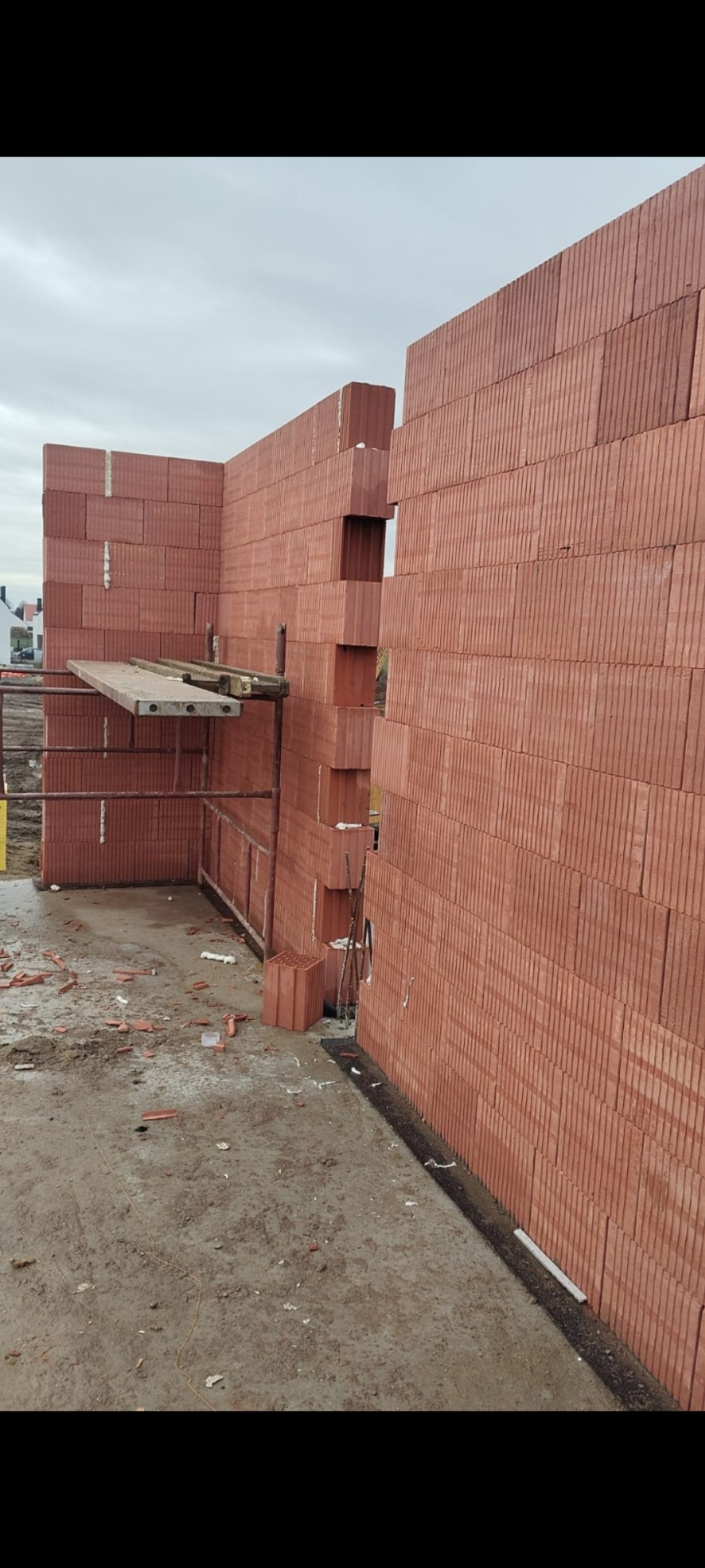 Wznoszone ściany domu z czerwonej cegły ceramicznej z widocznym rusztowaniem i fragmentami cegieł na ziemi.