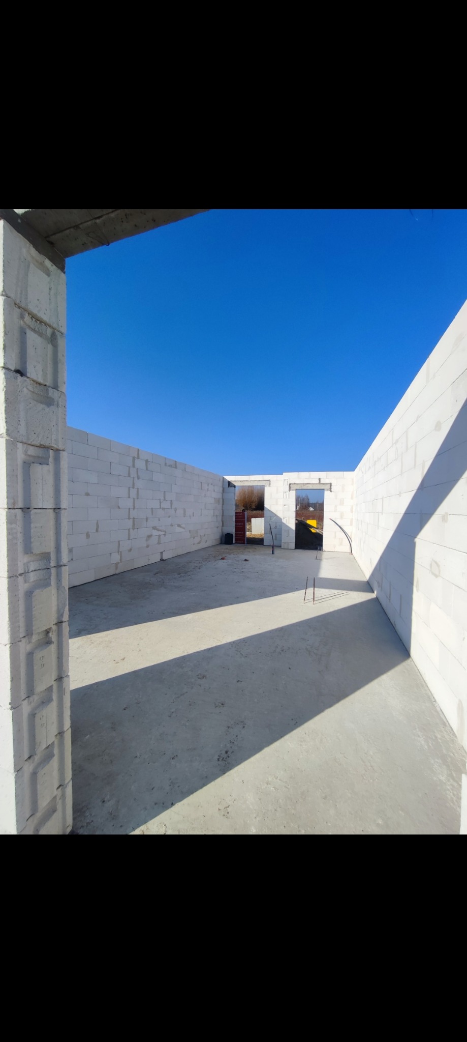 Wnętrze surowego budynku z białych bloczków, widać wylany betonowy fundament, niebo błękitne widoczne przez otwory okienne i drzwiowe, w tle widoczne drzewa.