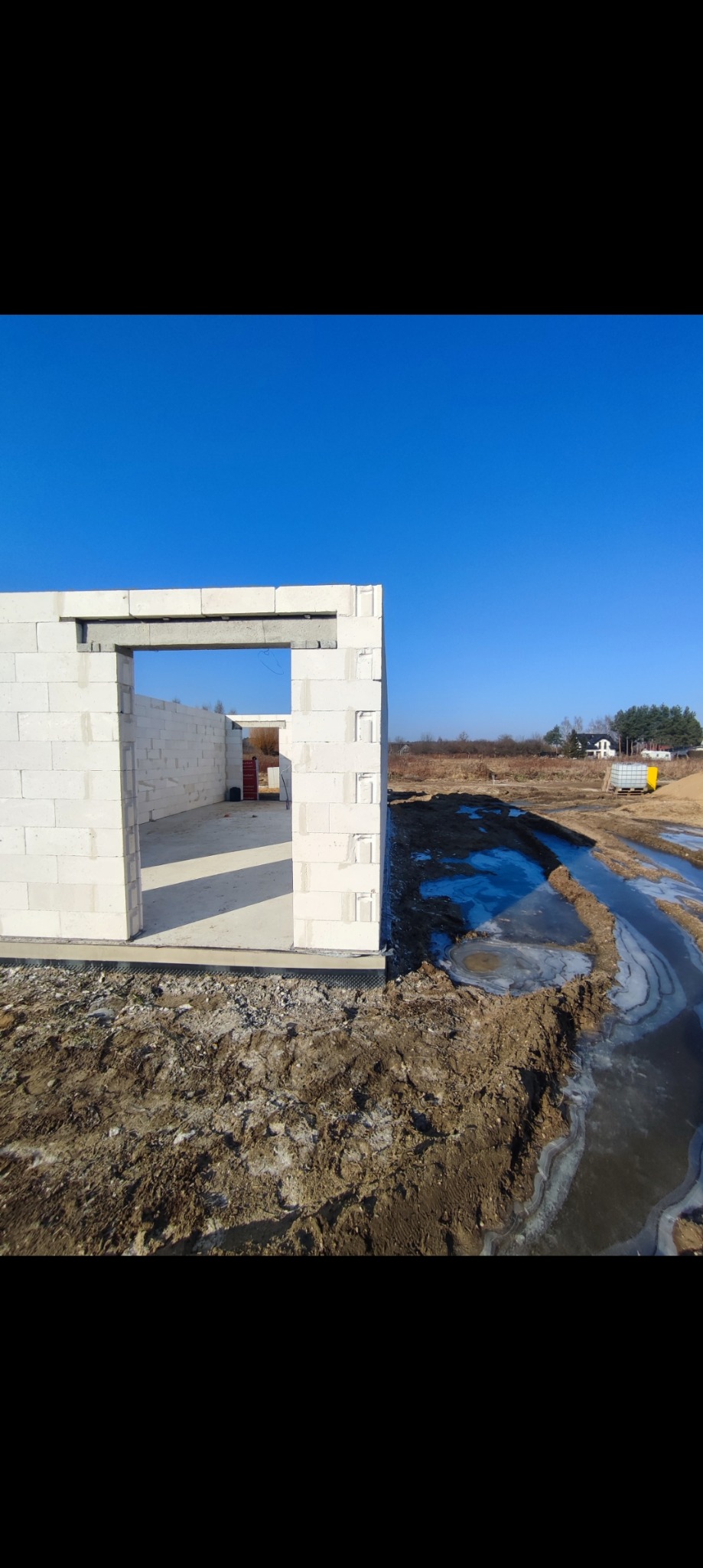 Wznoszone ściany z bloczków z betonu komórkowego na fundamencie, widoczne wstępne otwory okienne i drzwiowe, teren wokół w stanie surowym, częściowo zamarznięty.