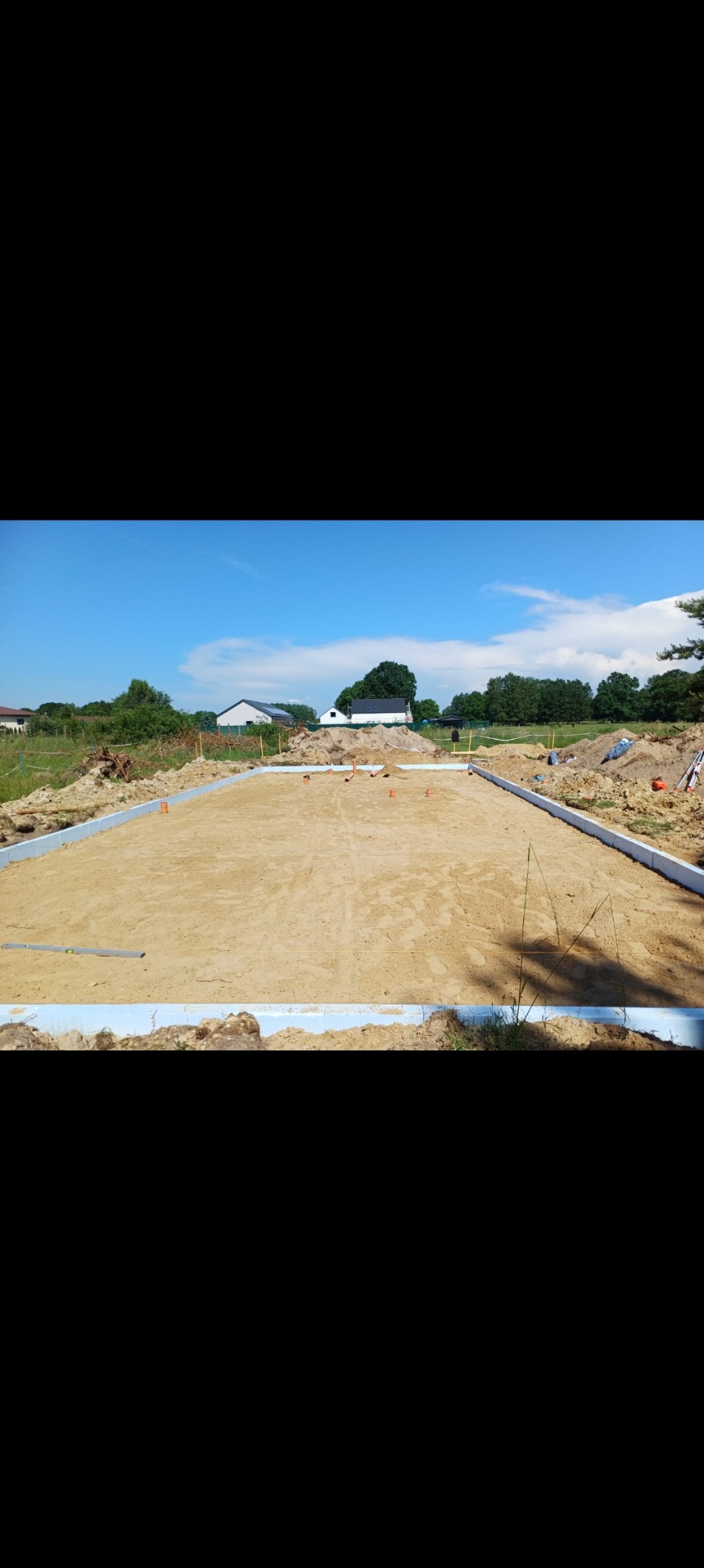 Przygotowany teren pod budowę domu z wyznaczonym obrysem fundamentów i izolacją przeciwwilgociową, widok na otwartą przestrzeń i zabudowania w oddali.