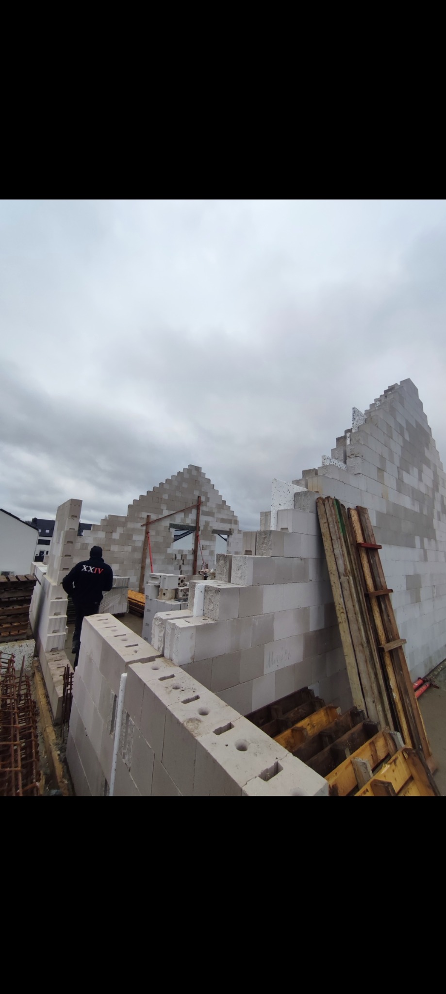 Wznoszenie ścian domu z bloczków betonu komórkowego, widoczny pracownik w czarnej bluzie z napisem 'XXIV', szalunki i zbrojenia w tle, pochmurne niebo.