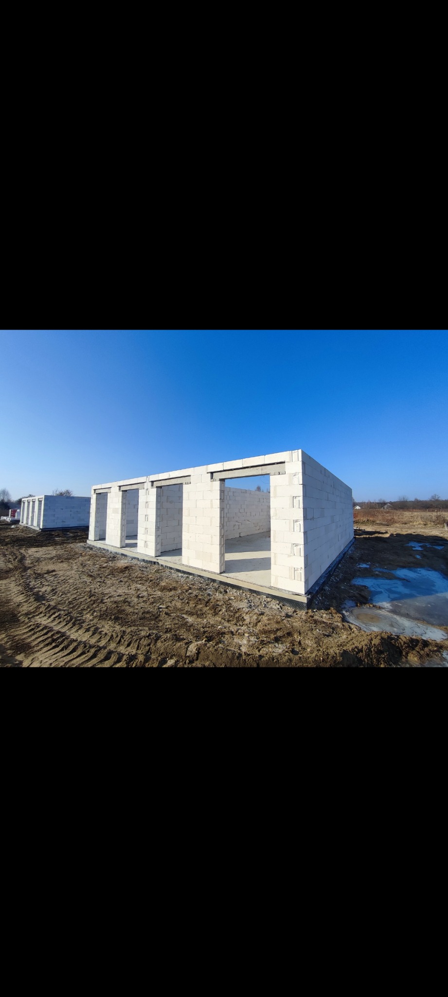 Wznoszenie ścian parterowego budynku z bloczków z betonu komórkowego, widoczne początki ścian i otwory okienne, na zewnątrz widoczny grunt i ślady opon.