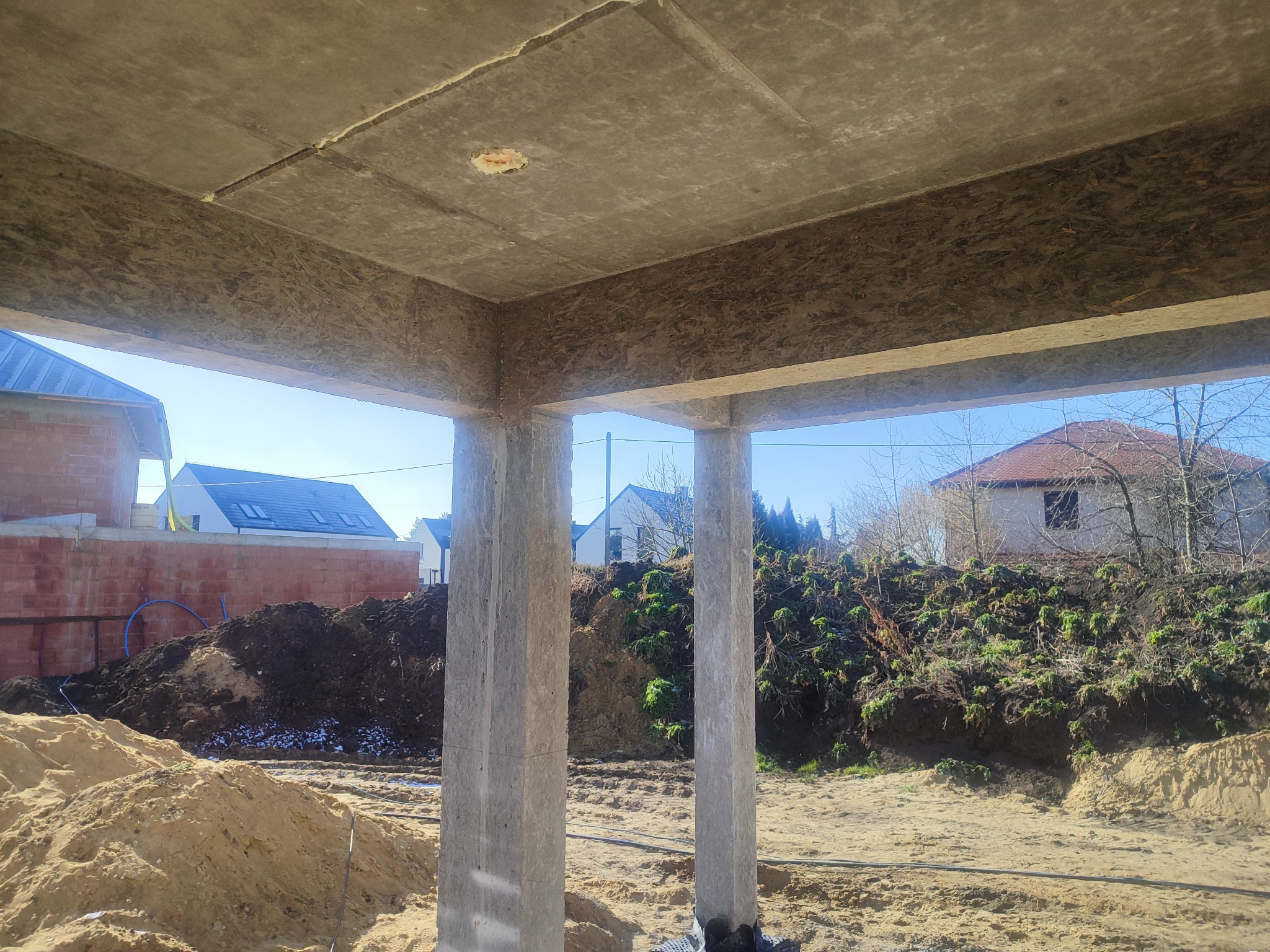 Wnętrze budynku w stanie surowym otwartym z widocznymi betonowymi słupami podtrzymującymi strop, w tle hałdy piasku i ziemi oraz budynki w różnym stadium budowy.