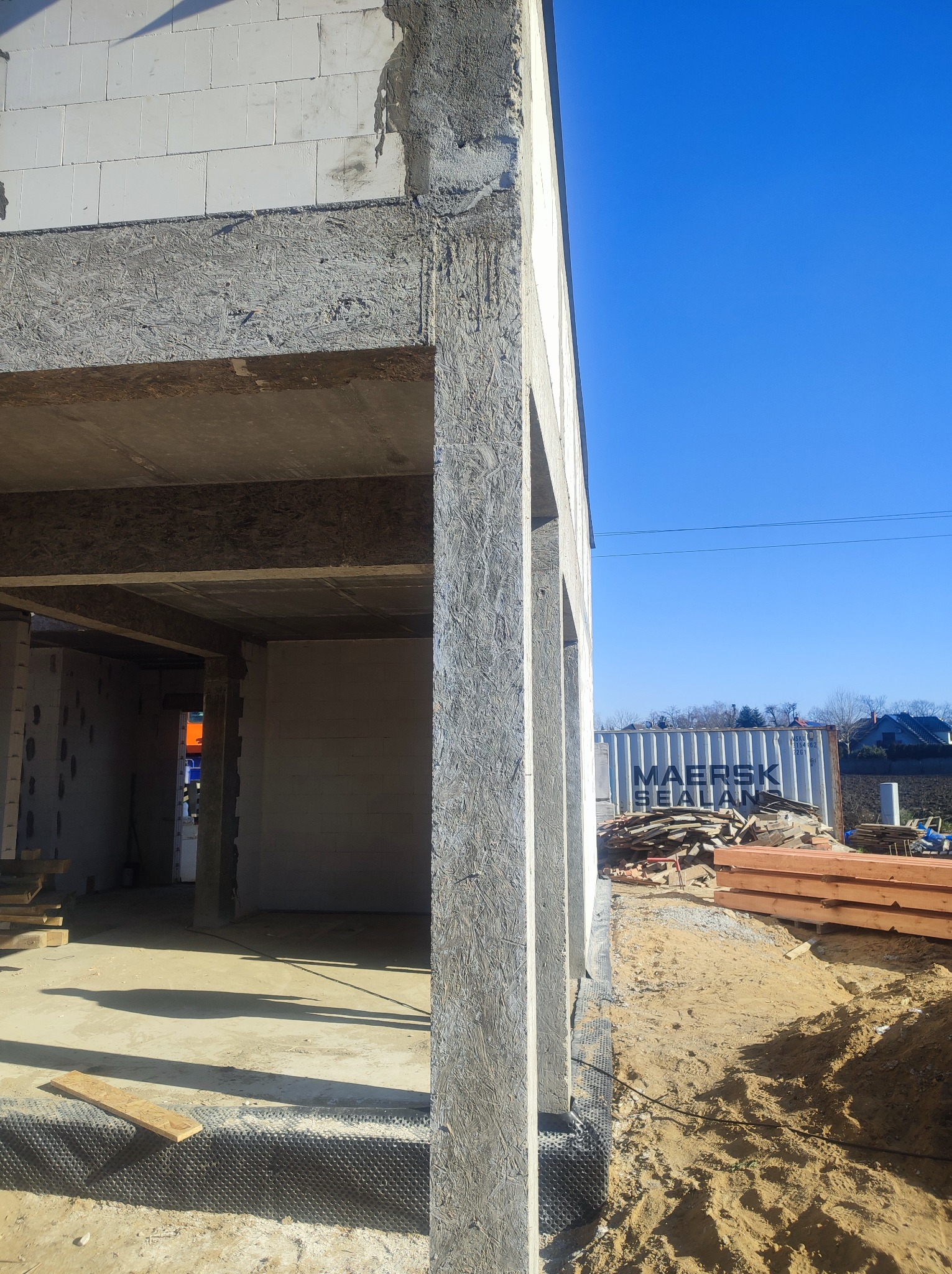 Surowy stan domu w trakcie budowy: ściany z betonu komórkowego, szalunki z płyty OSB, widoczna izolacja przeciwwilgociowa fundamentów, w tle kontener transportowy.