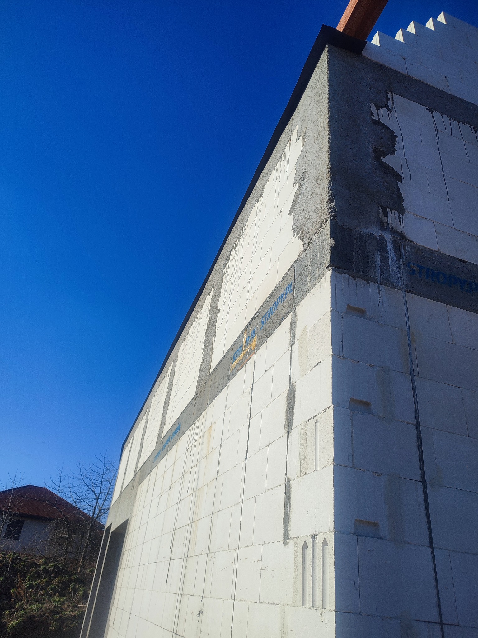 Ściana domu murowanego w trakcie budowy, widoczne białe bloczki, zbrojenie i fragment drewnianej konstrukcji dachu na tle błękitnego nieba.