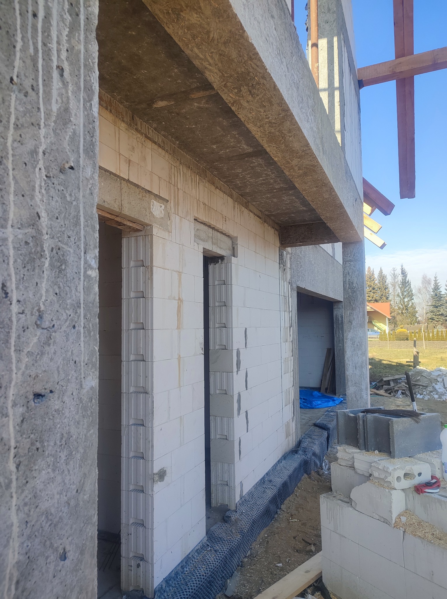 Wznoszenie ścian domu z betonu komórkowego, widoczne otwory okienne i drzwiowe, zabezpieczenie fundamentów folią kubełkową, w tle drzewa i fragment zabudowy jednorodzinnej.