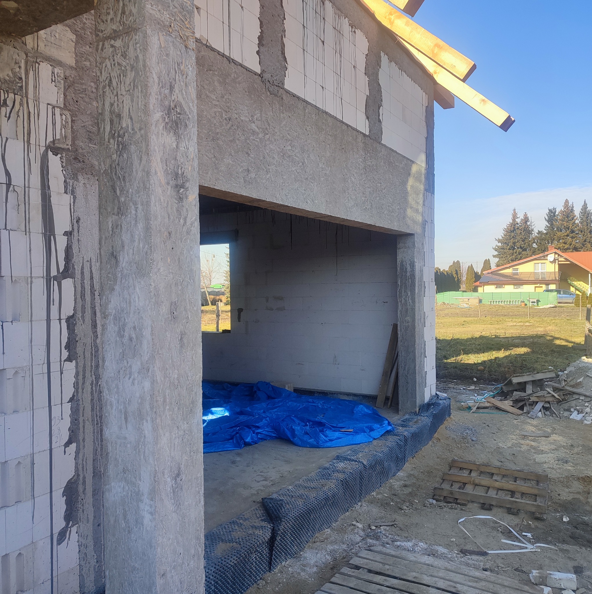 Wznoszenie ścian parteru z bloczków, widoczna izolacja przeciwwilgociowa fundamentów oraz niebieska plandeka zabezpieczająca podłoże wewnątrz budynku.