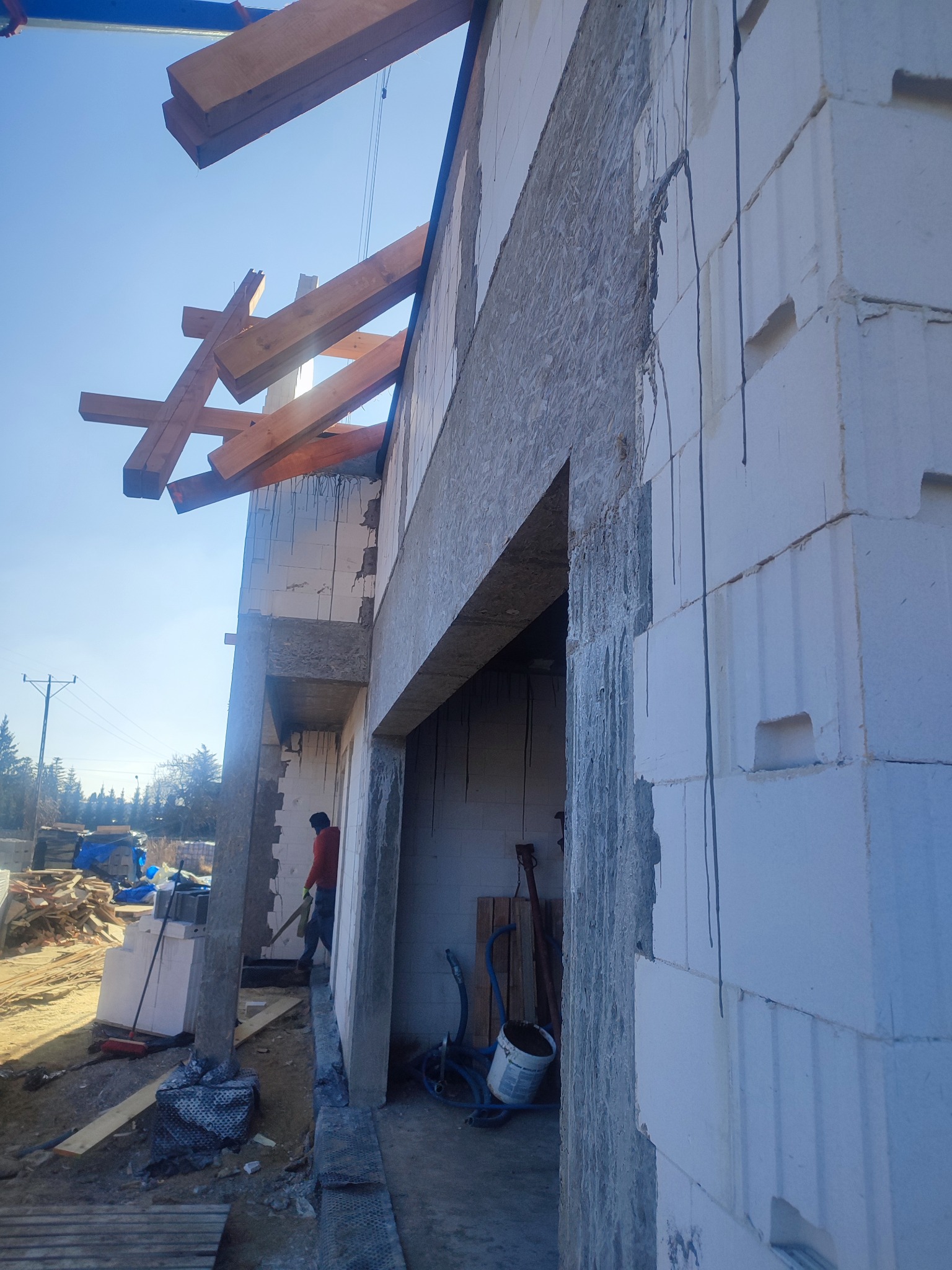 Wznoszenie ścian domu z bloczków betonu komórkowego, widoczne drewniane elementy konstrukcyjne dachu i pracownik w czerwonej kurtce.