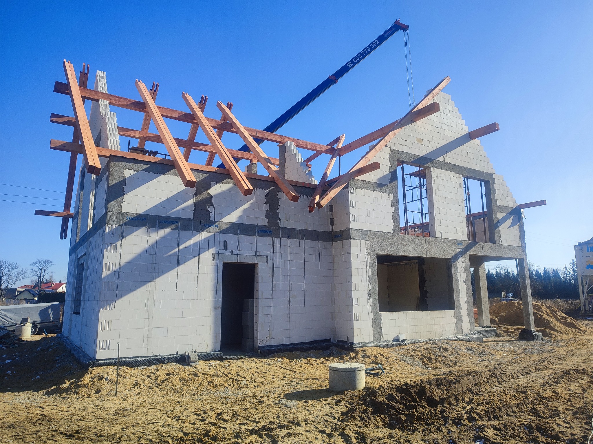 Wznoszenie dwukondygnacyjnego domu z białych bloczków, widoczne drewniane elementy konstrukcji dachu i pracujący żuraw budowlany na tle błękitnego nieba.