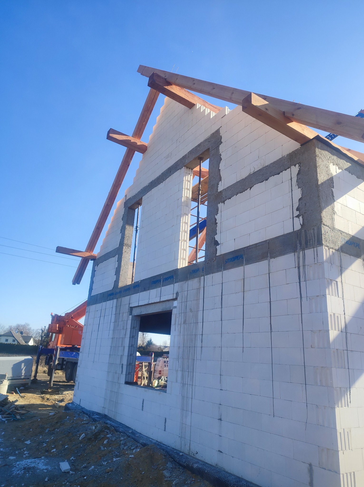 Wznoszenie ścian domu z białych bloczków, widoczne drewniane elementy konstrukcji dachu oraz dźwig w tle.