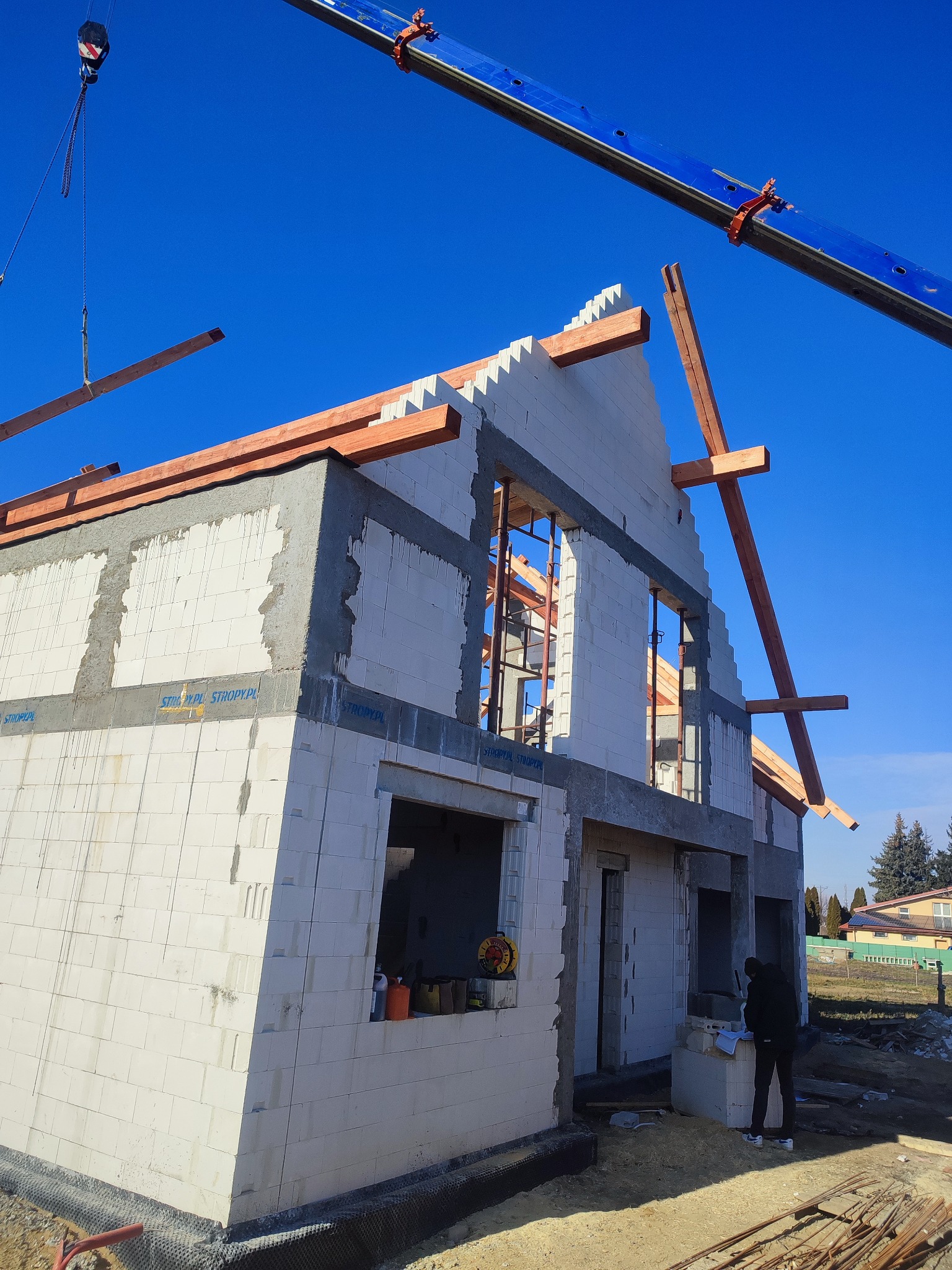 Wznoszenie dwukondygnacyjnego domu z białych bloczków, widoczny dźwig podający drewniane elementy konstrukcji dachu, na pierwszym planie osoba przeglądająca dokumenty.