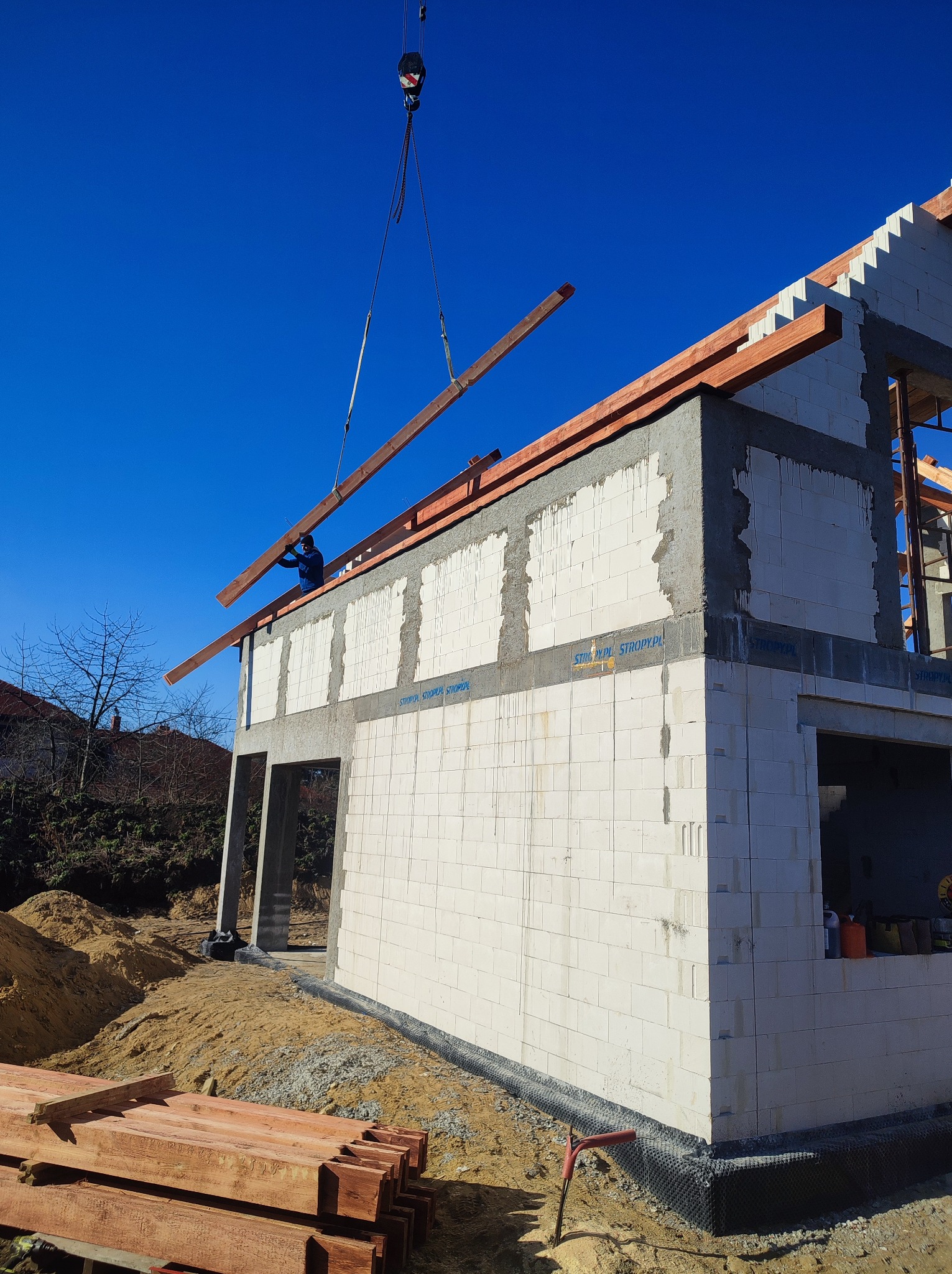 Wznoszenie ścian domu z betonu komórkowego; dźwig podnosi drewniany element konstrukcji dachu, widoczna izolacja przeciwwilgociowa fundamentów.