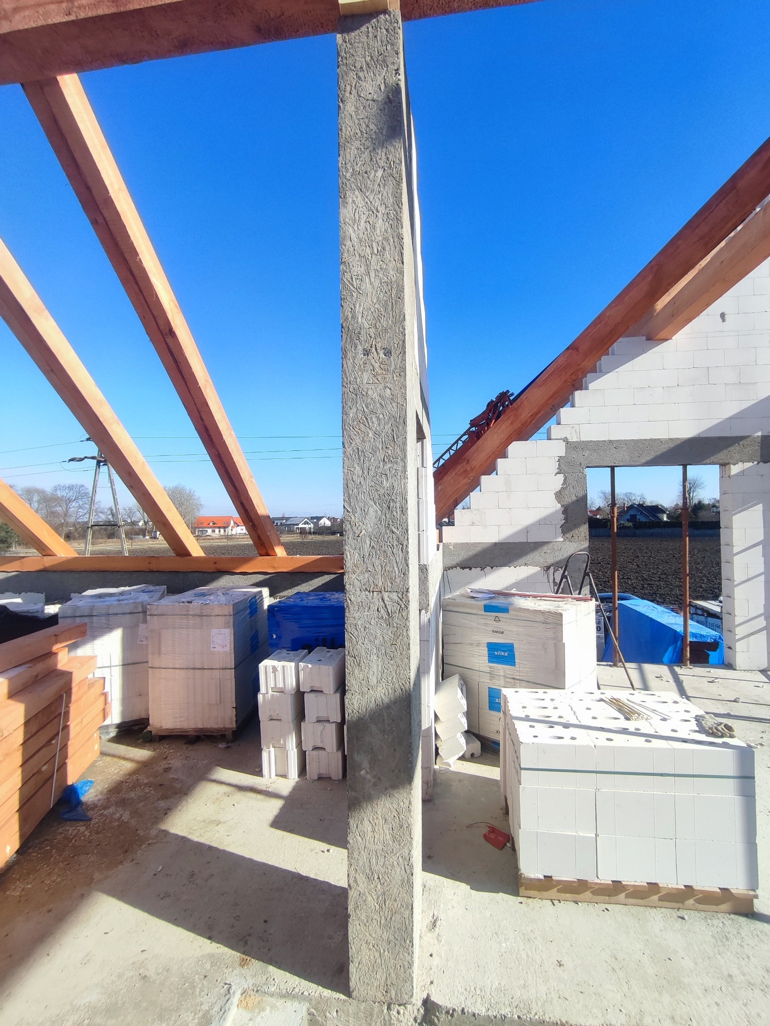 Wnętrze domu w trakcie budowy z widocznymi ścianami z bloczków, drewnianą konstrukcją dachu, ułożonymi paletami z materiałami budowlanymi i błękitnym niebem w tle.