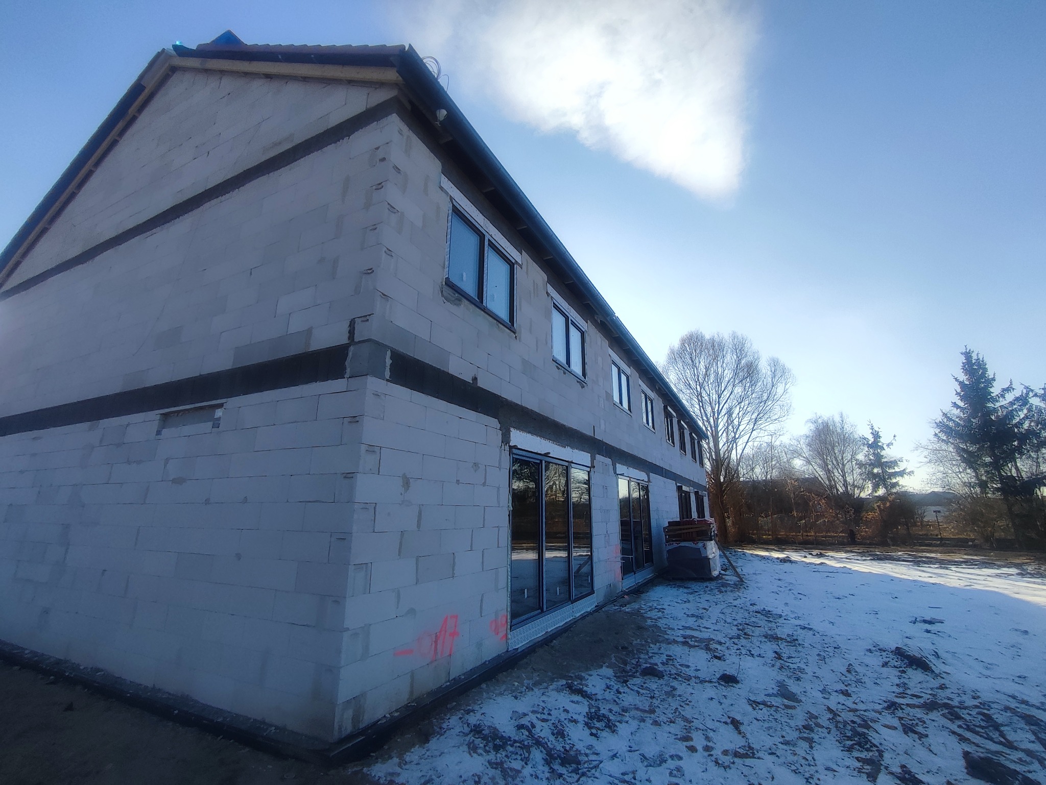 Surowy budynek z betonu komórkowego w stanie surowym zamkniętym, z zamontowanymi oknami i drzwiami balkonowymi, częściowo ośnieżony teren wokół.