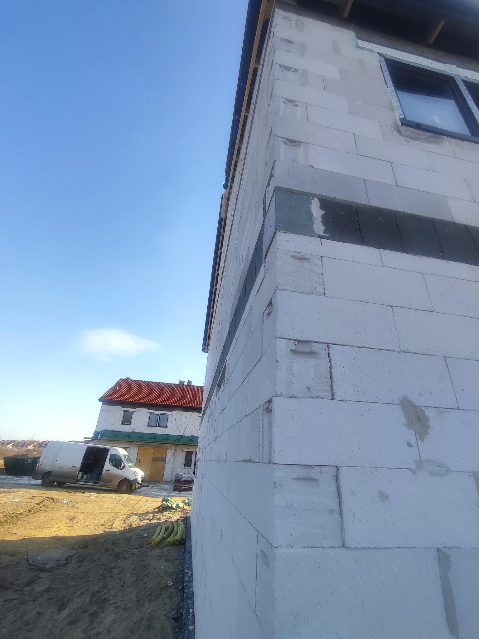 Wznoszona ściana domu z białych bloczków, widoczne szare wzmocnienia, w tle budynek z czerwoną dachówką i biały dostawczy samochód.