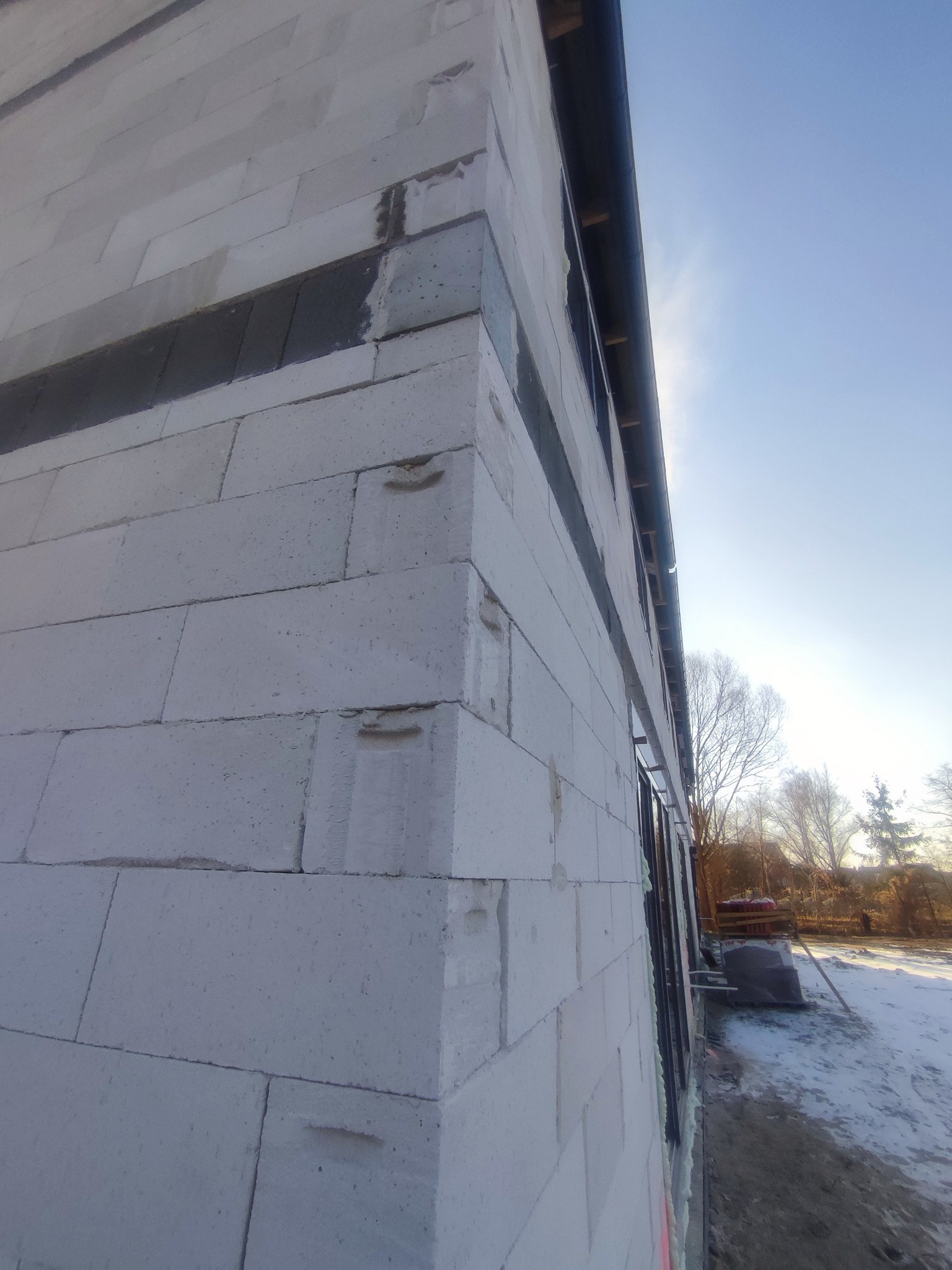 Niedokończony narożnik domu murowanego z betonu komórkowego, widoczne ślady po uchwytach w bloczkach, czarne pasy zaprawy, okno z pianką montażową, zaśnieżone otoczenie w tle.