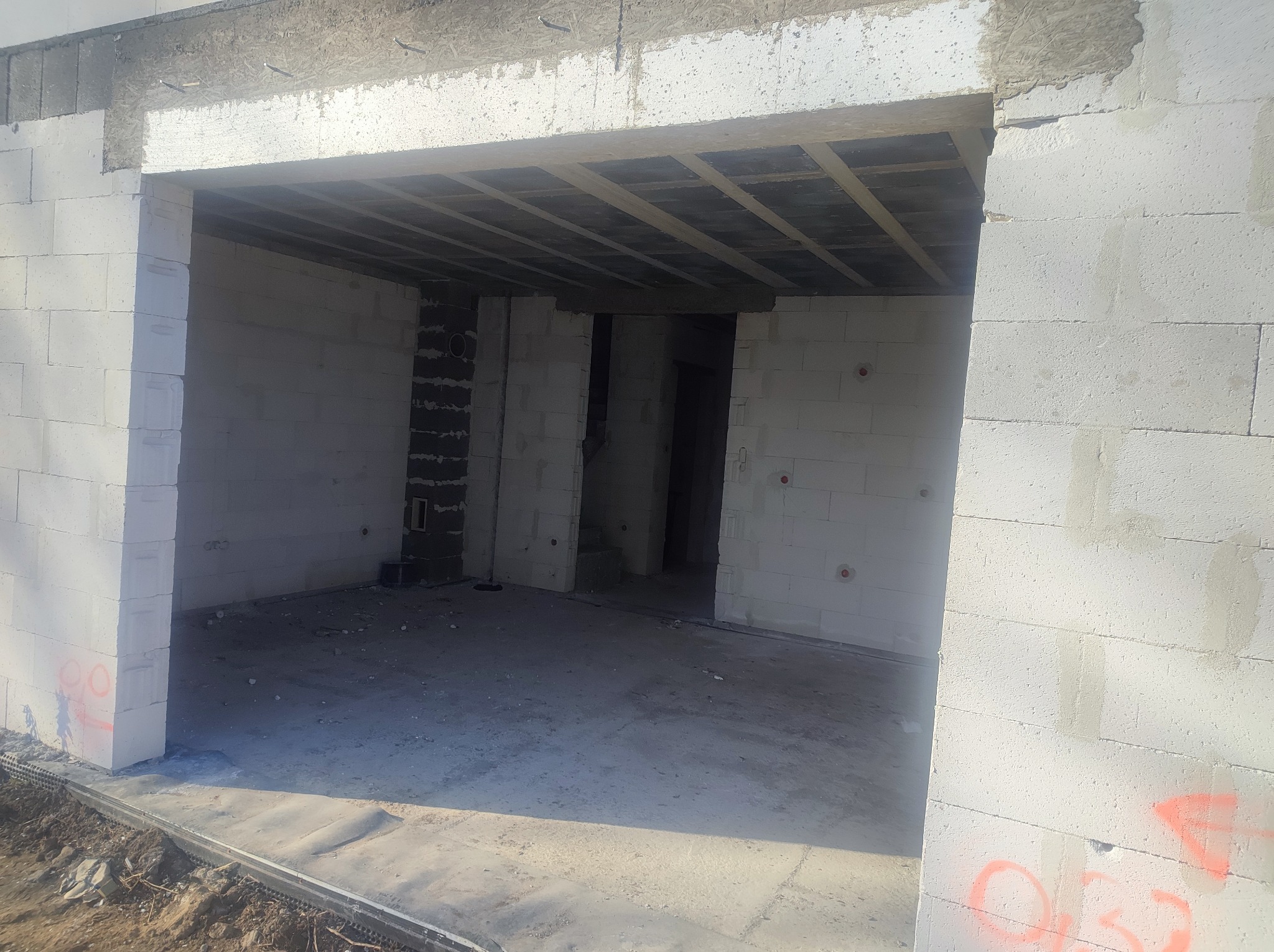 Surowy stan wnętrza domu w trakcie budowy, ściany z białych bloczków, widoczne zbrojenie nad otworem drzwiowym i sufit z drewnianych belek.