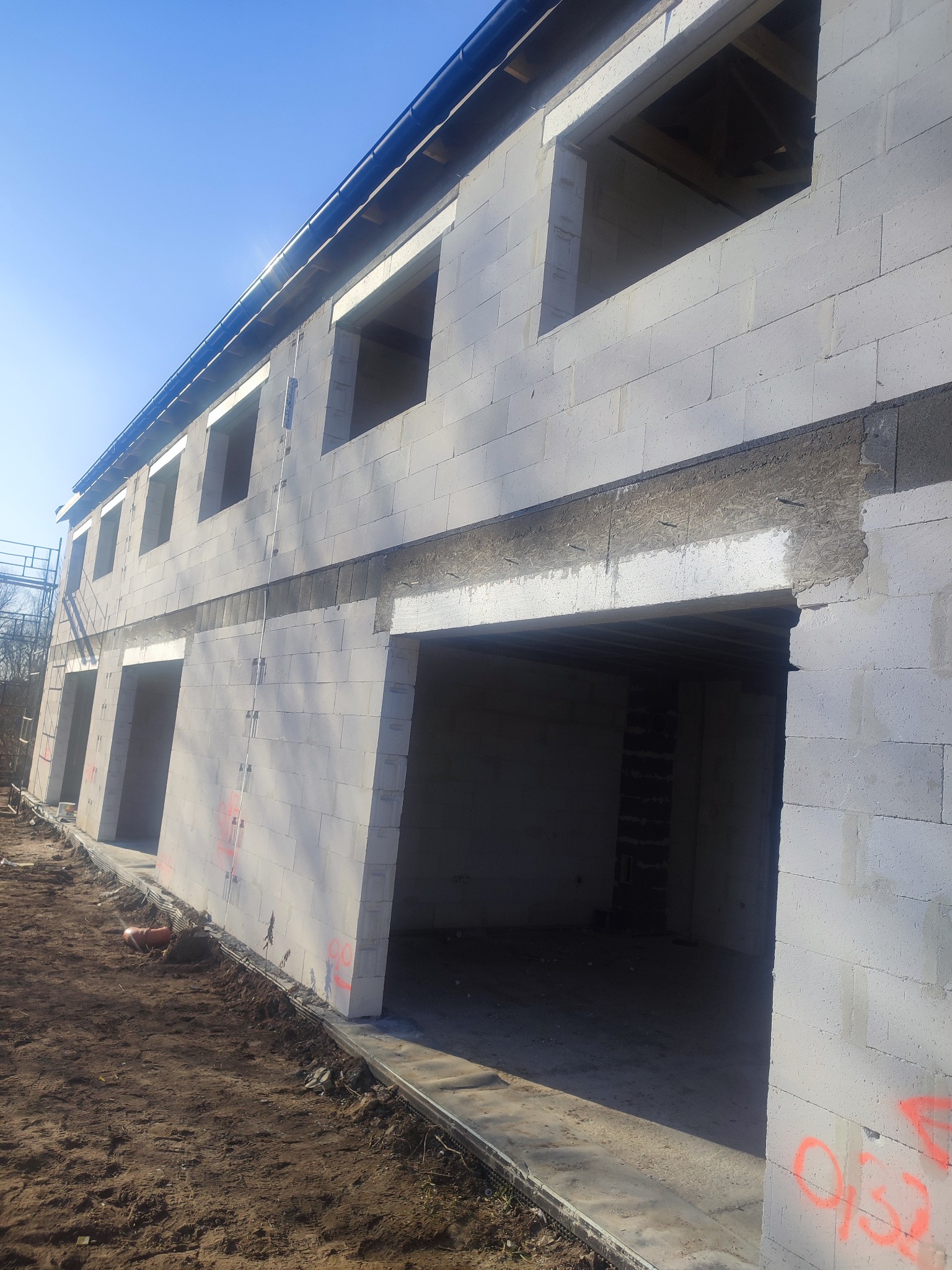 Wznoszenie ścian dwukondygnacyjnego budynku z bloczków betonu komórkowego, widoczne otwory okienne i drzwiowe, częściowo ocieplony fundament, w tle drzewa.