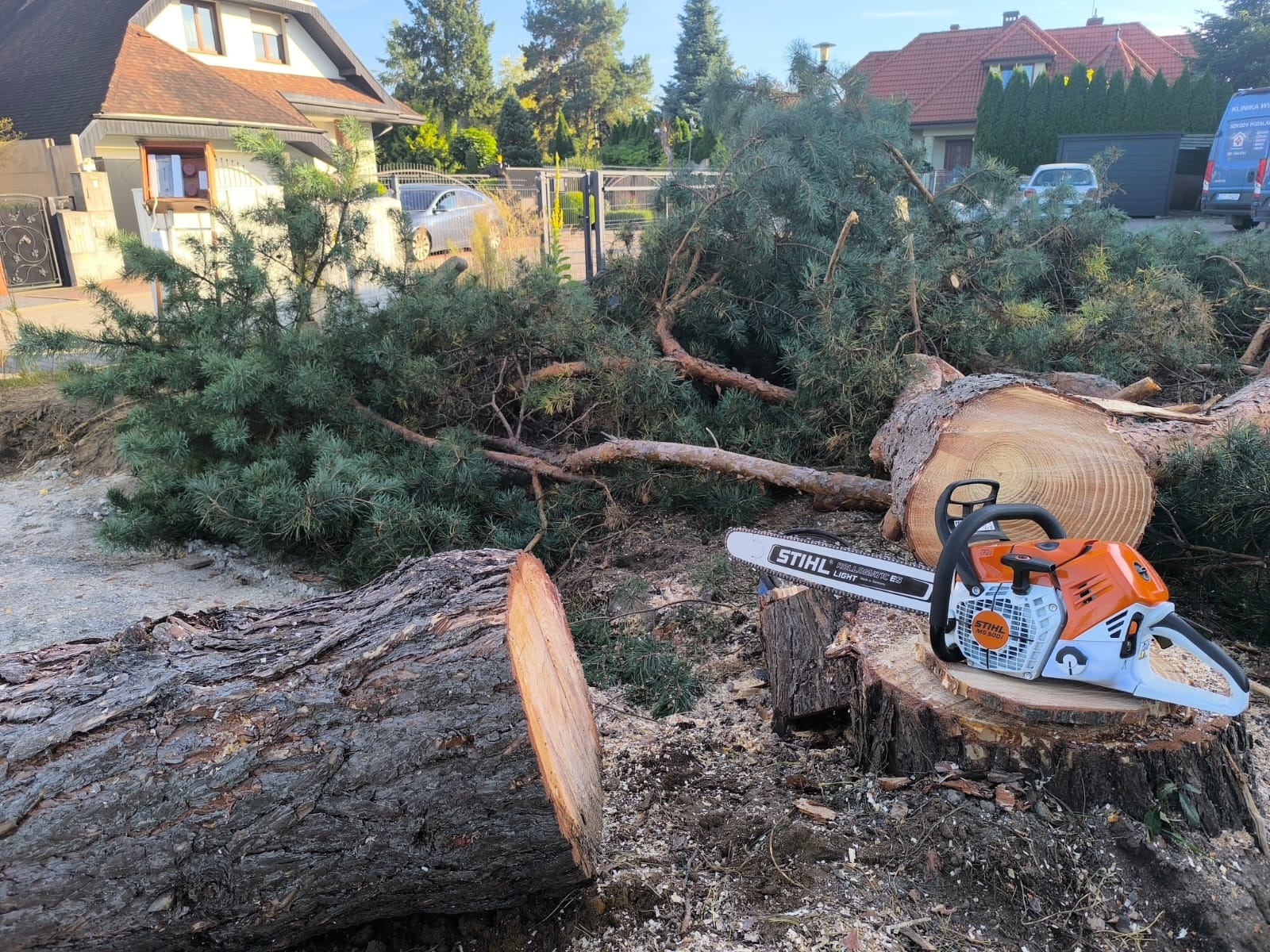 Ścięte gałęzie i pnie drzewa leżące na ziemi, obok piła spalinowa Stihl. W tle dom i samochód. Widok po wycince drzewa.