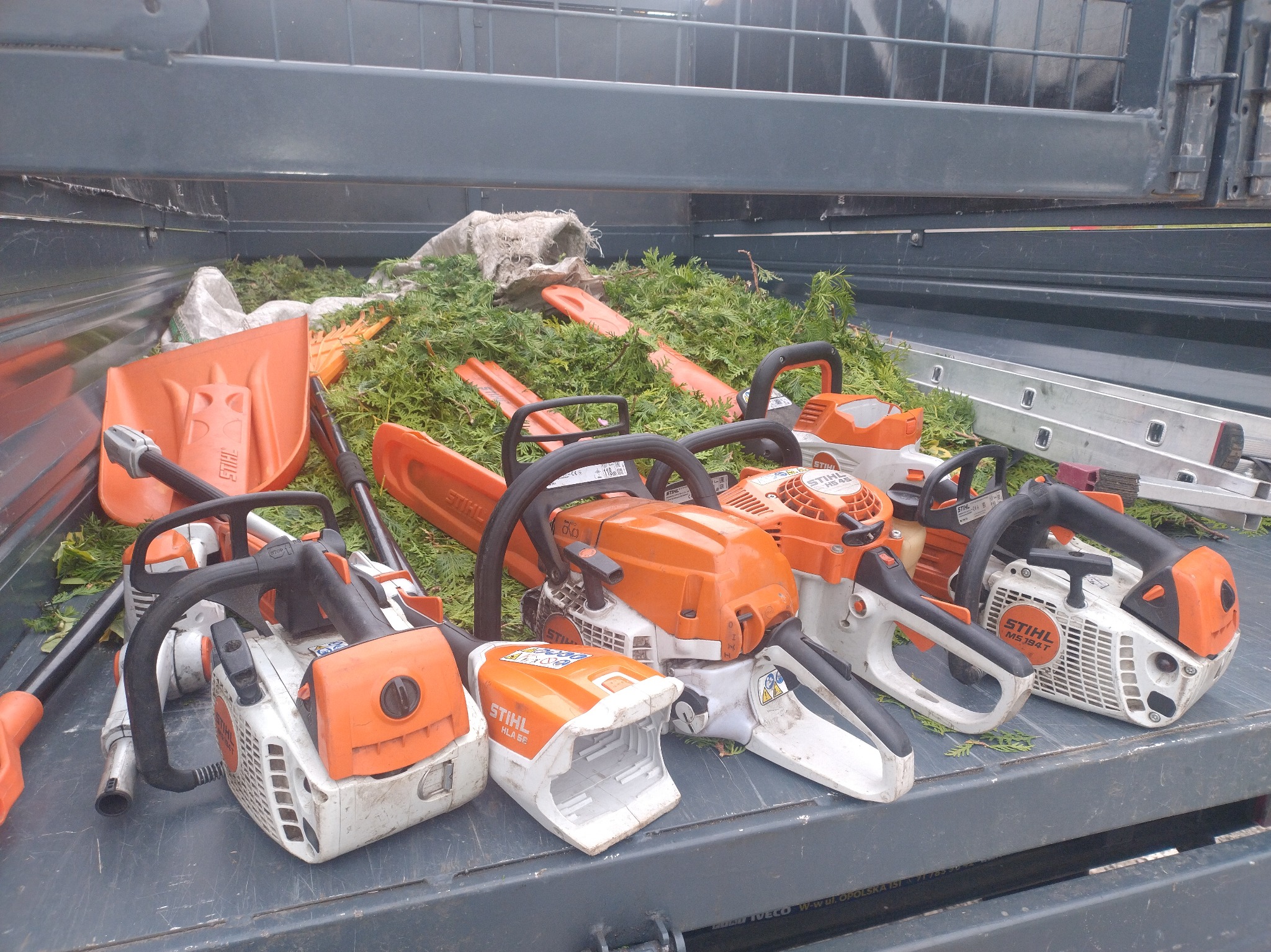 Przyczepa z narzędziami ogrodniczymi: pilarki Stihl, łopata i drabina. Widoczne ścięte gałęzie. Sprzęt gotowy do pracy.