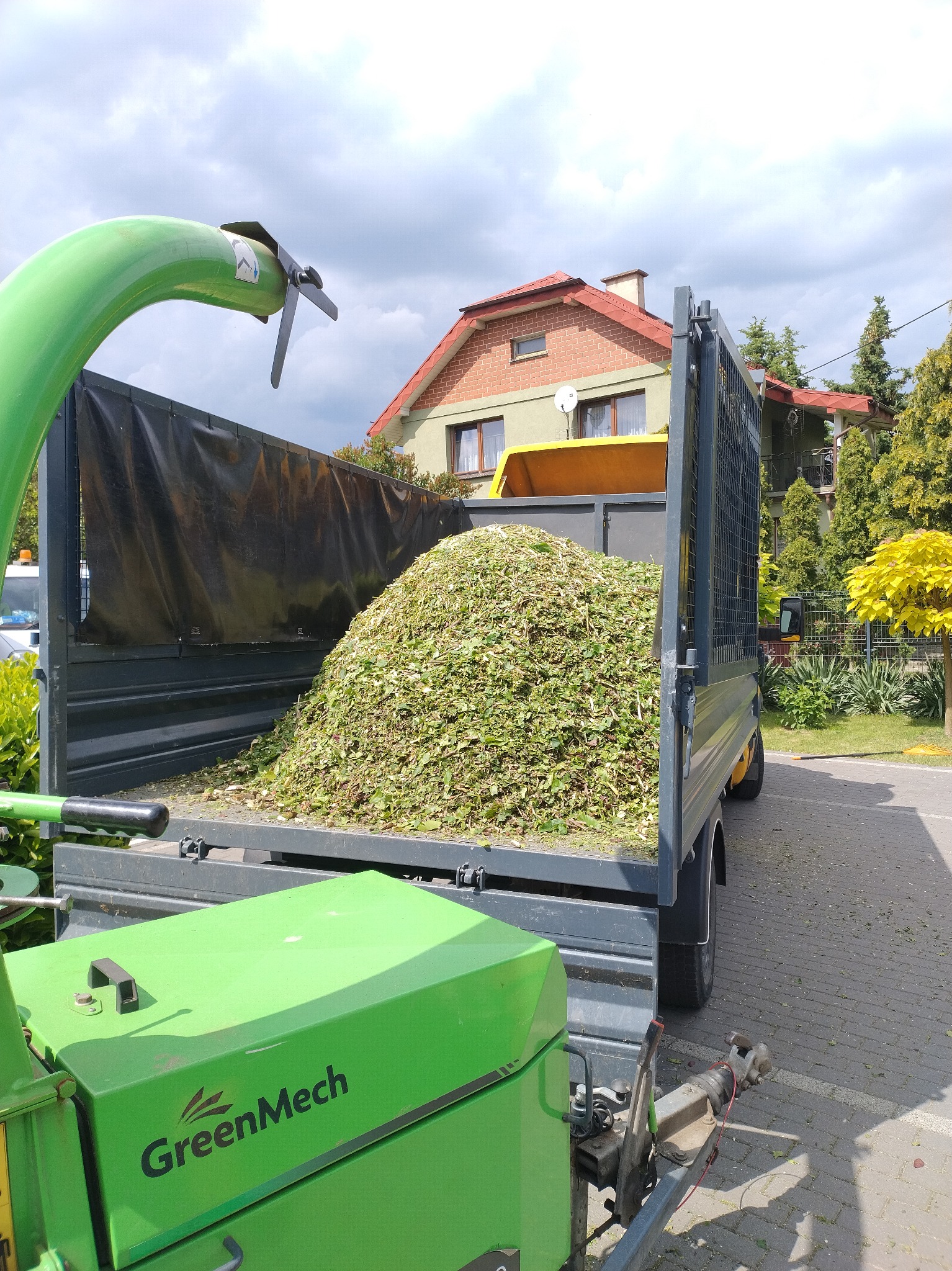 Ciężarówka wypełniona po brzegi świeżo rozdrobnionymi gałęziami i liśćmi, z widoczną zieleń maszyną GreenMech w tle, dom i drzewa w tle.