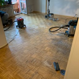 Volodymyr Pasichnyk - Parkiet w jodełkę podczas renowacji: maszyna do cyklinowania, odkurzacz przemysłowy, widoczne ślady po cyklinowaniu. W tle balkon z roślinami.