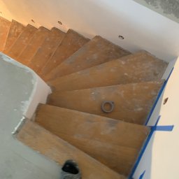 Volodymyr Pasichnyk - Drewniane schody spiralne w trakcie renowacji, zabezpieczone taśmą malarską, widok z góry. Na schodach leży rolka taśmy. W tle ściany przygotowane do malowania.
