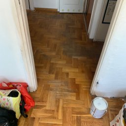 Volodymyr Pasichnyk - Parkiet ułożony w jodełkę, częściowo wycyklinowany, z widocznymi śladami użytkowania i puszką farby w rogu. Widok z góry na fragment podłogi w trakcie renowacji.