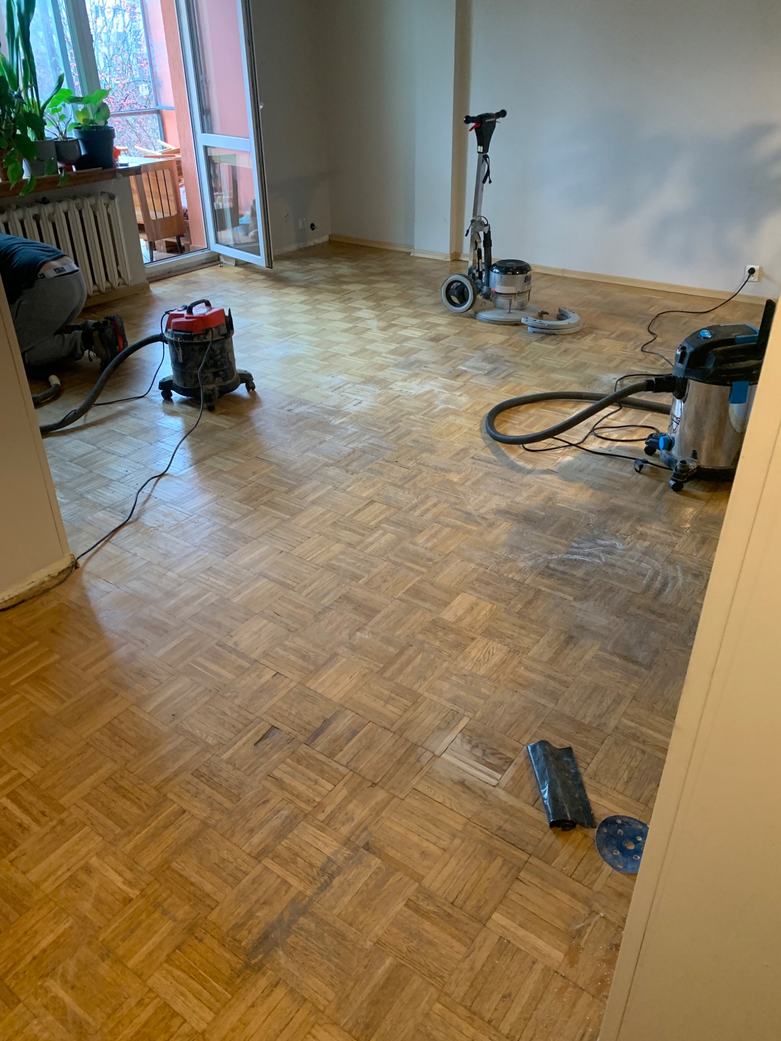 Parkiet w jodełkę podczas renowacji: maszyna do cyklinowania, odkurzacz przemysłowy, widoczne ślady po cyklinowaniu. W tle balkon z roślinami.