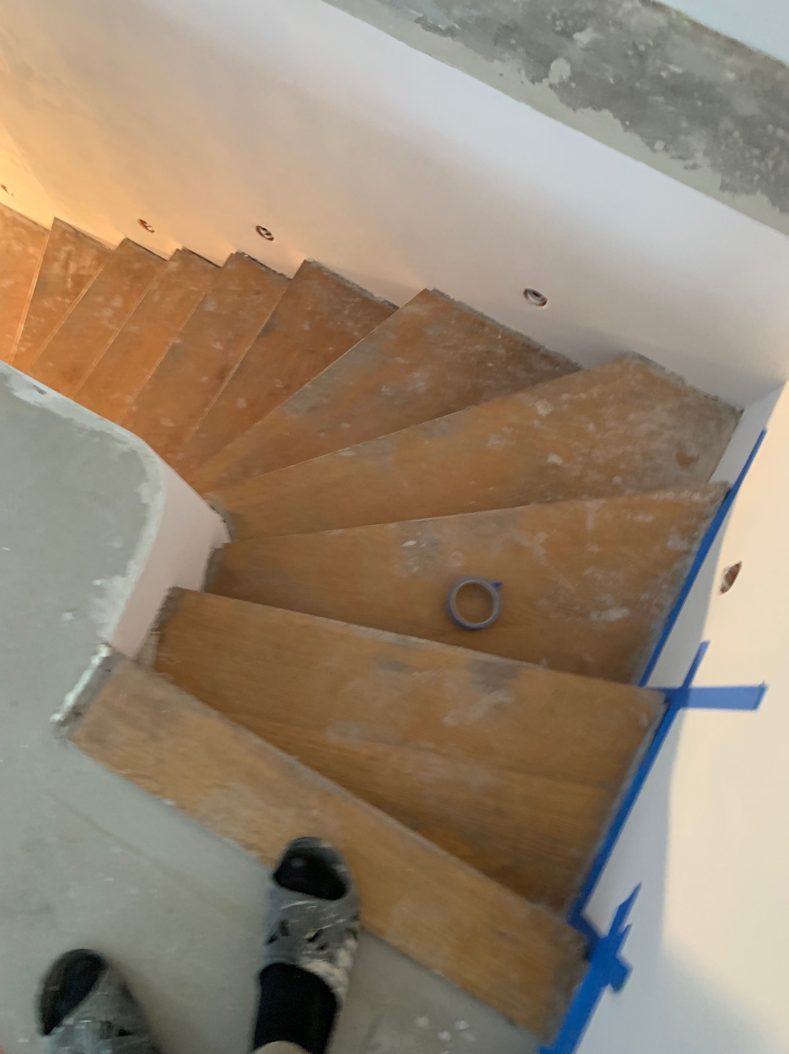 Drewniane schody spiralne w trakcie renowacji, zabezpieczone taśmą malarską, widok z góry. Na schodach leży rolka taśmy. W tle ściany przygotowane do malowania.